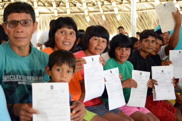 Indígenas Waimiri Atroari sentados, lado a lado, segurando registros de nascimento dentro de uma maloca na comunidade.