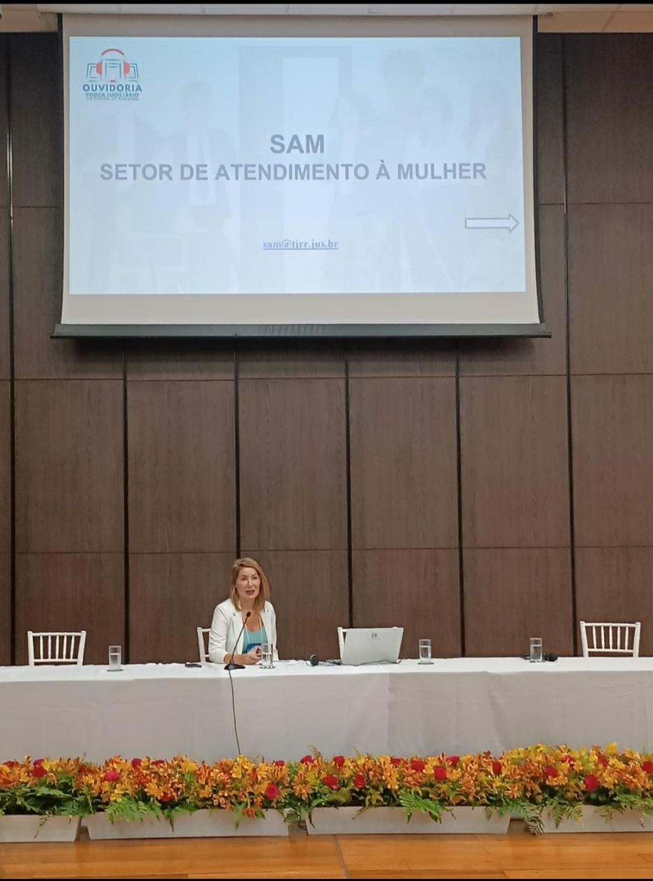 foto colorida da mesa de apresentações nela está uma mulher e acima estava um slide escrito SAM SETOR DE ATENDIMENTO A MULHER