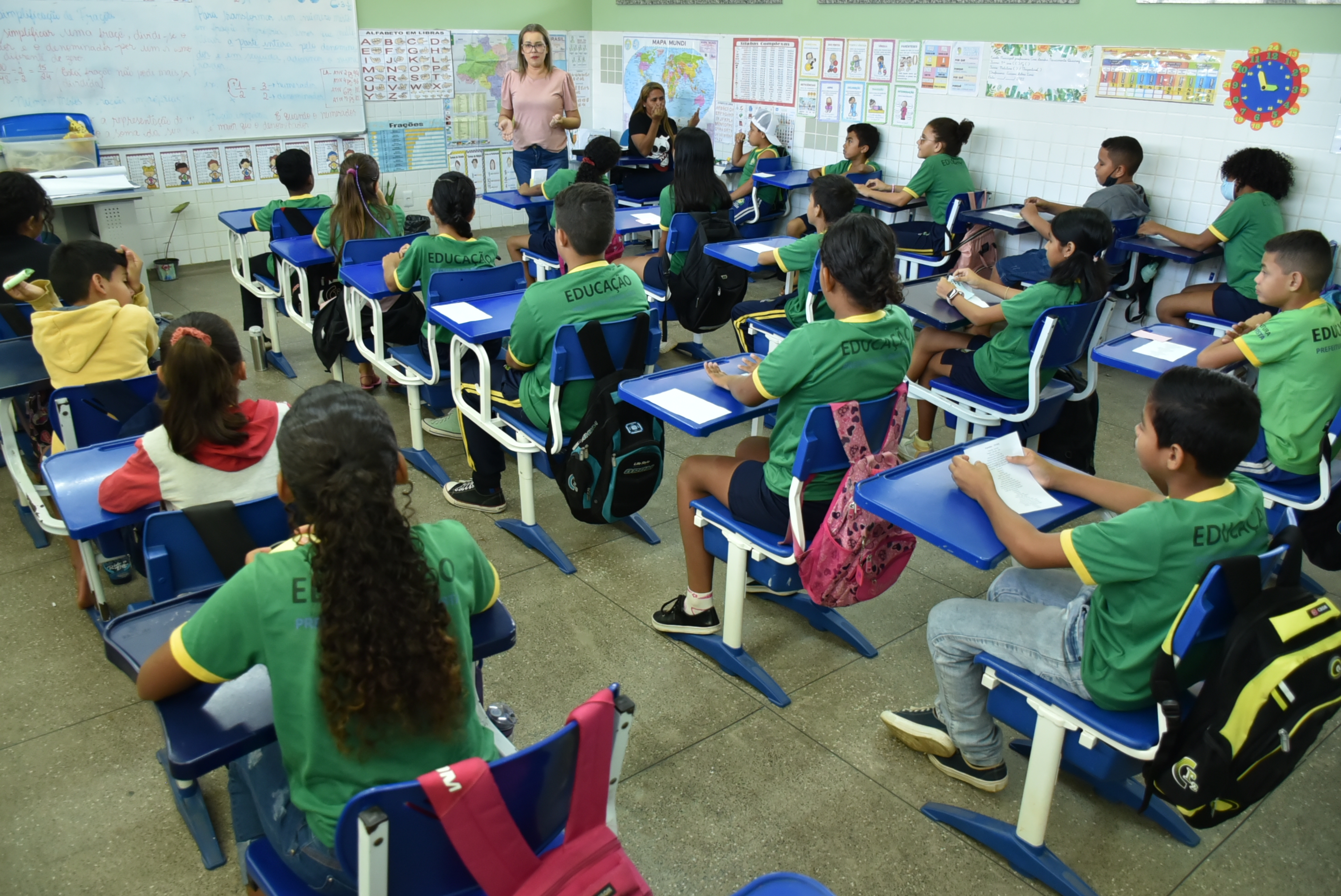 Foto colorida mostra aproximadamente 20 crianças sentadas em suas cadeiras na sala de aula prestando atenção na professora