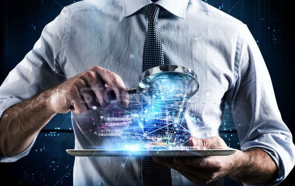 Imagem do torso de um homem engravatado que segura uma lupa visualizando luzes saindo de um tablet que está sendo segurado pela mão esquerda do mesmo.