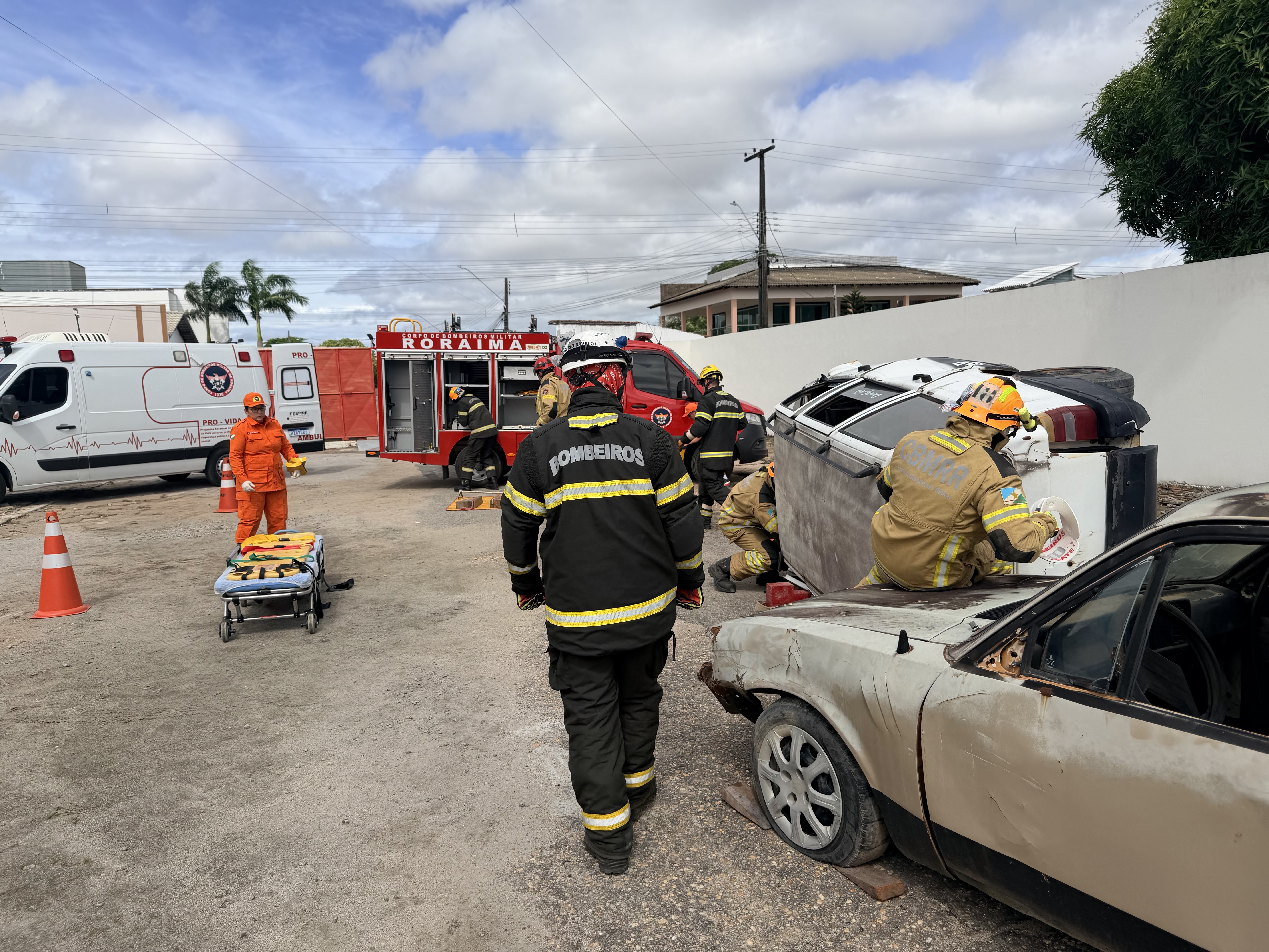 Vista ampla da área de treinamento. Ao centro, um bombeiro de costas com o uniforme escrito “BOMBEIROS”. Ao fundo, uma ambulância e um caminhão de combate a incêndio vermelho escrito “RORAIMA”. À direita, bombeiros trabalham em um carro branco capotado. Vista ampla da área de treinamento. Ao centro, um bombeiro de costas com o uniforme escrito “BOMBEIROS”. Ao fundo, uma ambulância e um caminhão de combate a incêndio vermelho escrito “RORAIMA”. À direita, bombeiros trabalham em um carro branco capotado.