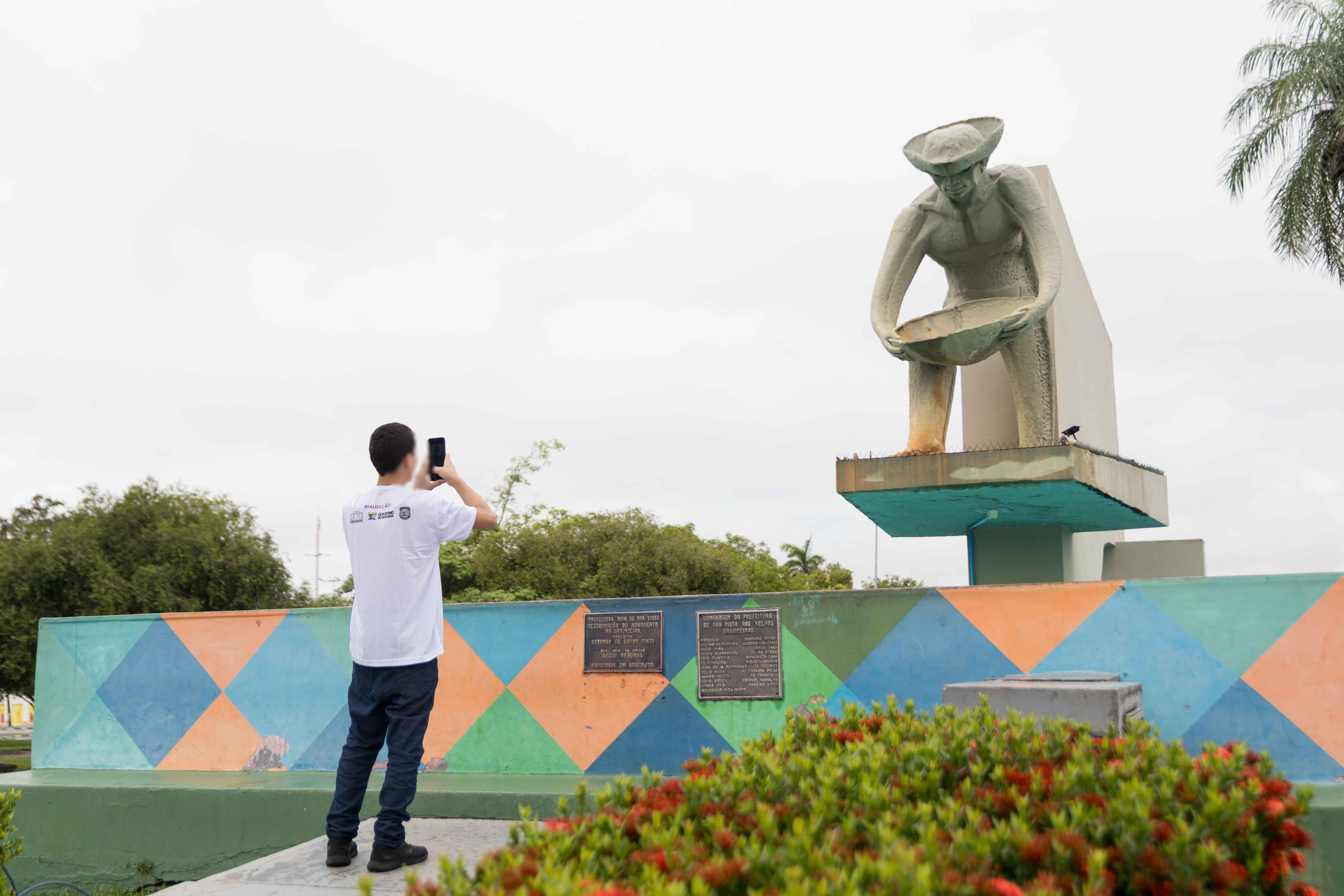 Foto colorida contém adolescente de costas fotografando o Monumento do Garimpeiro, localizado no Centro da Capital Boa Vista, Roraima. 