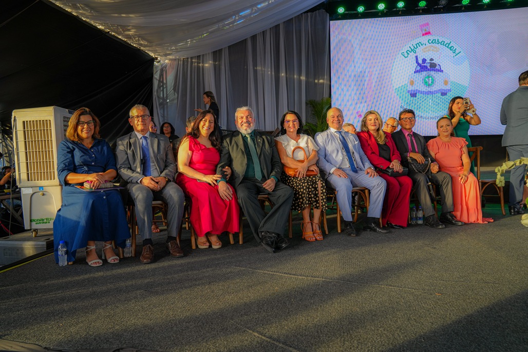 Grupo de nove pessoas sorrindo e sentadas em cadeiras, vestindo roupas formais e coloridas, em um evento com iluminação em um palco ao fundo. Grupo de nove pessoas sorrindo e sentadas em cadeiras, vestindo roupas formais e coloridas, em um evento com iluminação em um palco ao fundo.
