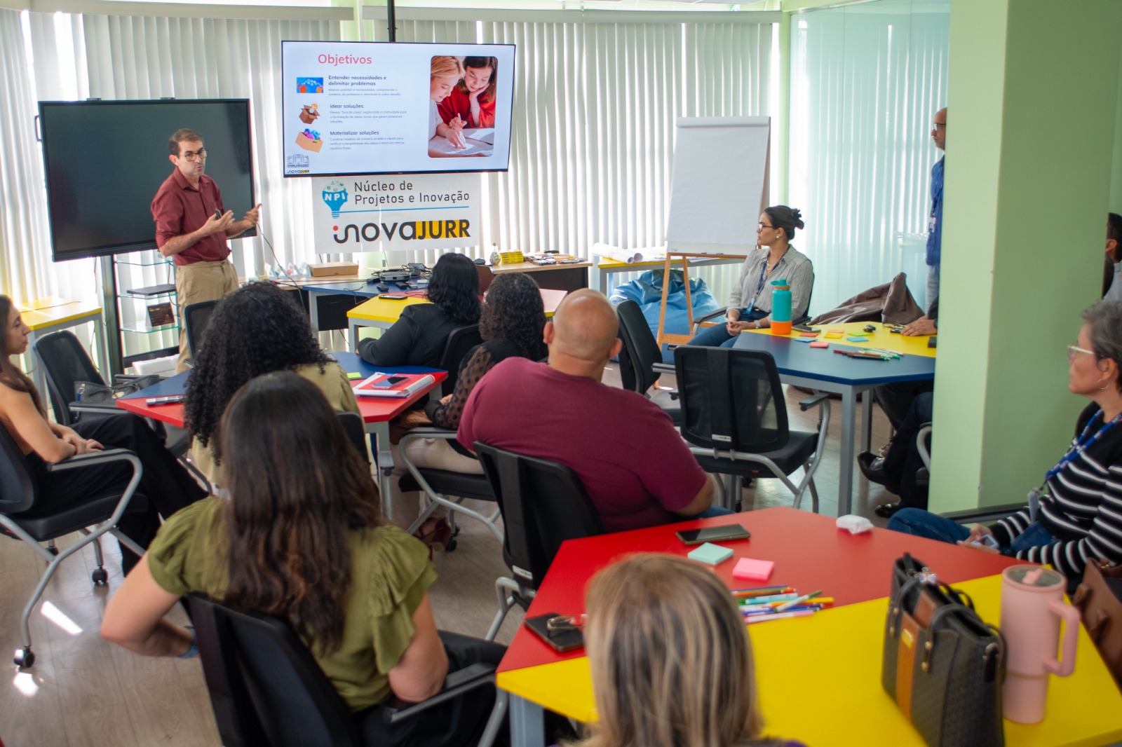 Foto colorida mostra uma sala de reunião com pessoas sentadas ao redor de mesas coloridas, assistindo a uma apresentação. Um homem em pé explica o conteúdo exibido em uma tela sobre objetivos de inovação.