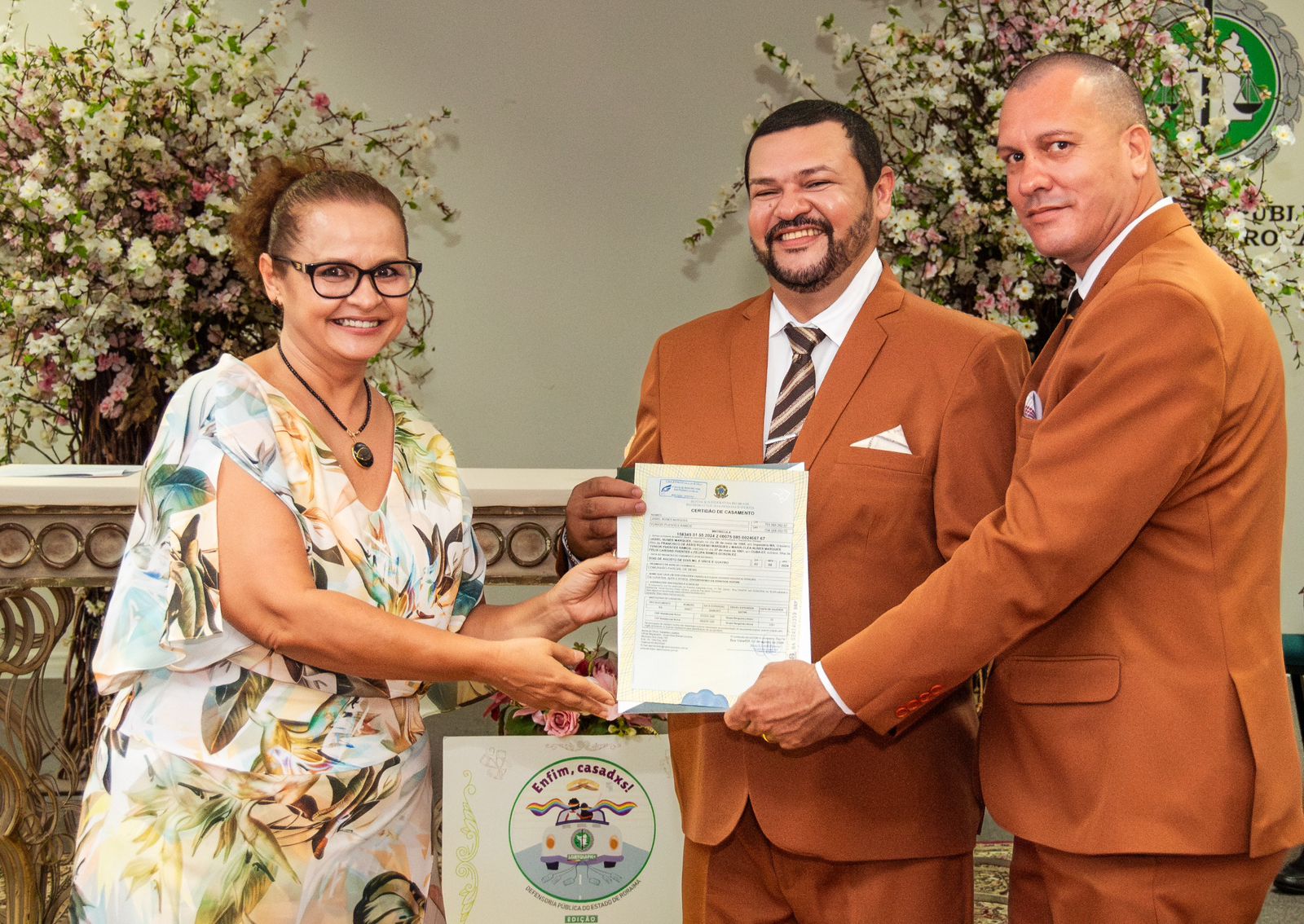 foto colorida de um casal de homens junto a juíza graciette sotto maior pousando para  a fotografia enquanto seguram o papel de oficialização de união