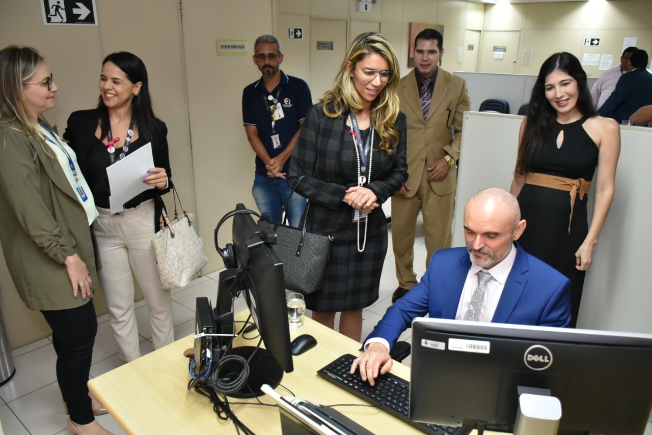 Imagem colorida em formato retangular mostra o juiz coordenador dos Juizados Especiais, Air Marin digitando em um computador com duas telas postas sobre uma mesa retangular de cor marfim, ao seu lado encontra-se a a diretora de relações institucionais e gerente do departamento jurídico da Roraima Energia, Sarassele Freire, juntamente com um grupo de pessoas, umas conversando e outras observando a ação do Juiz.