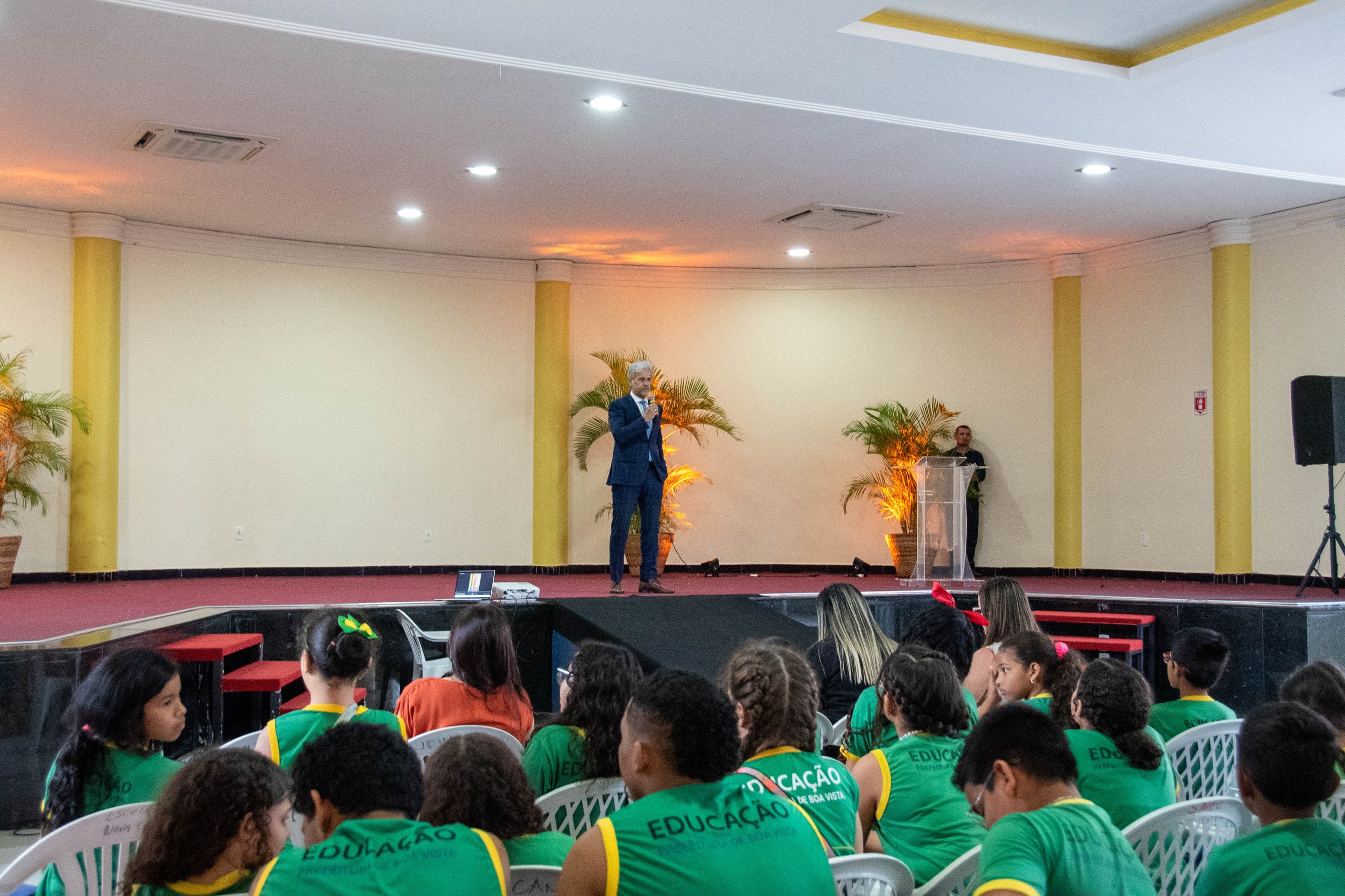 foto colorida do juiz titular do segundo juizado de Violência Doméstica e Familiar, representando a Cevid/TJRR, Jaime Plá, falando ao microfone em um auditório cheio de crianças da escola da prefeitura sentadas