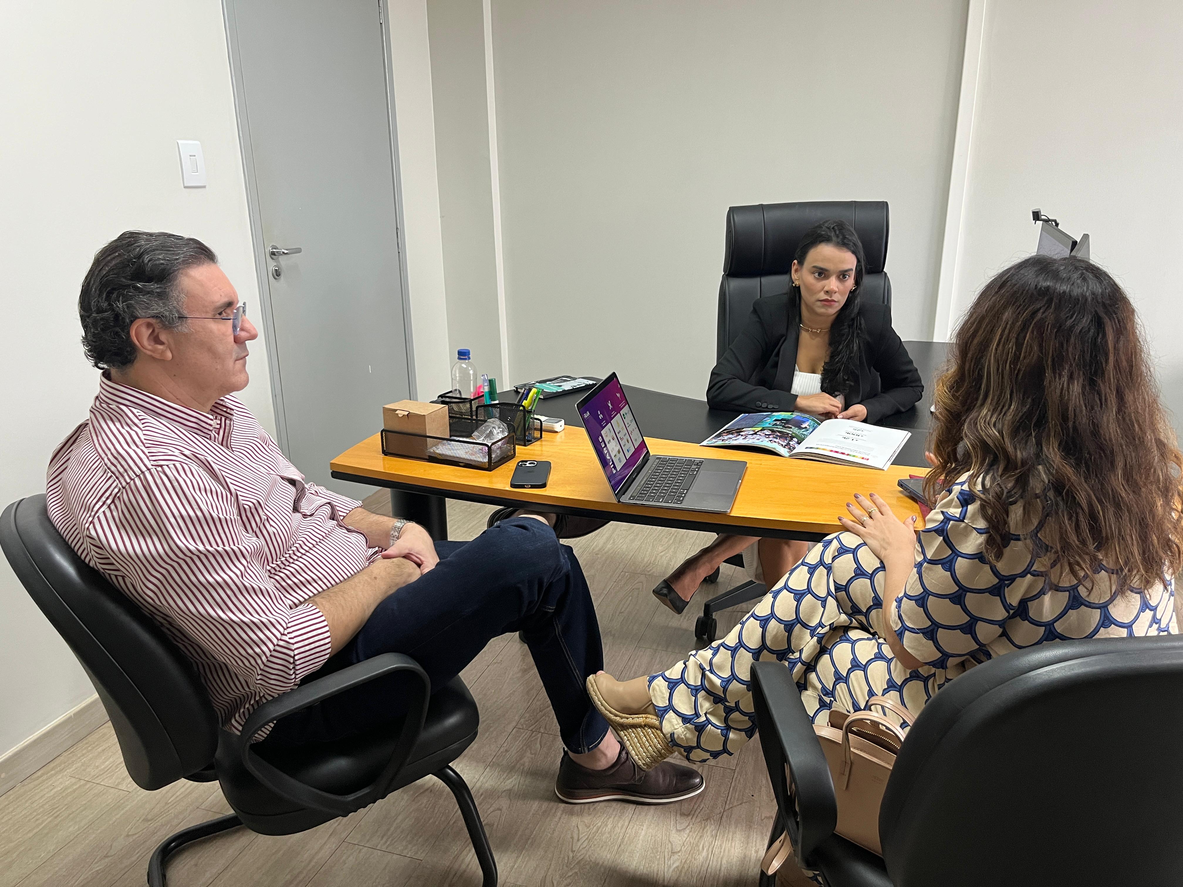 imagem colorida mostra três pessoas conversando em volta de uma mesa de reunião, Sendo duas mulheres e um homem vestidos de trajes sociais. Em cima da mesa se encontra notbook, papéis, e itens de escritório. 