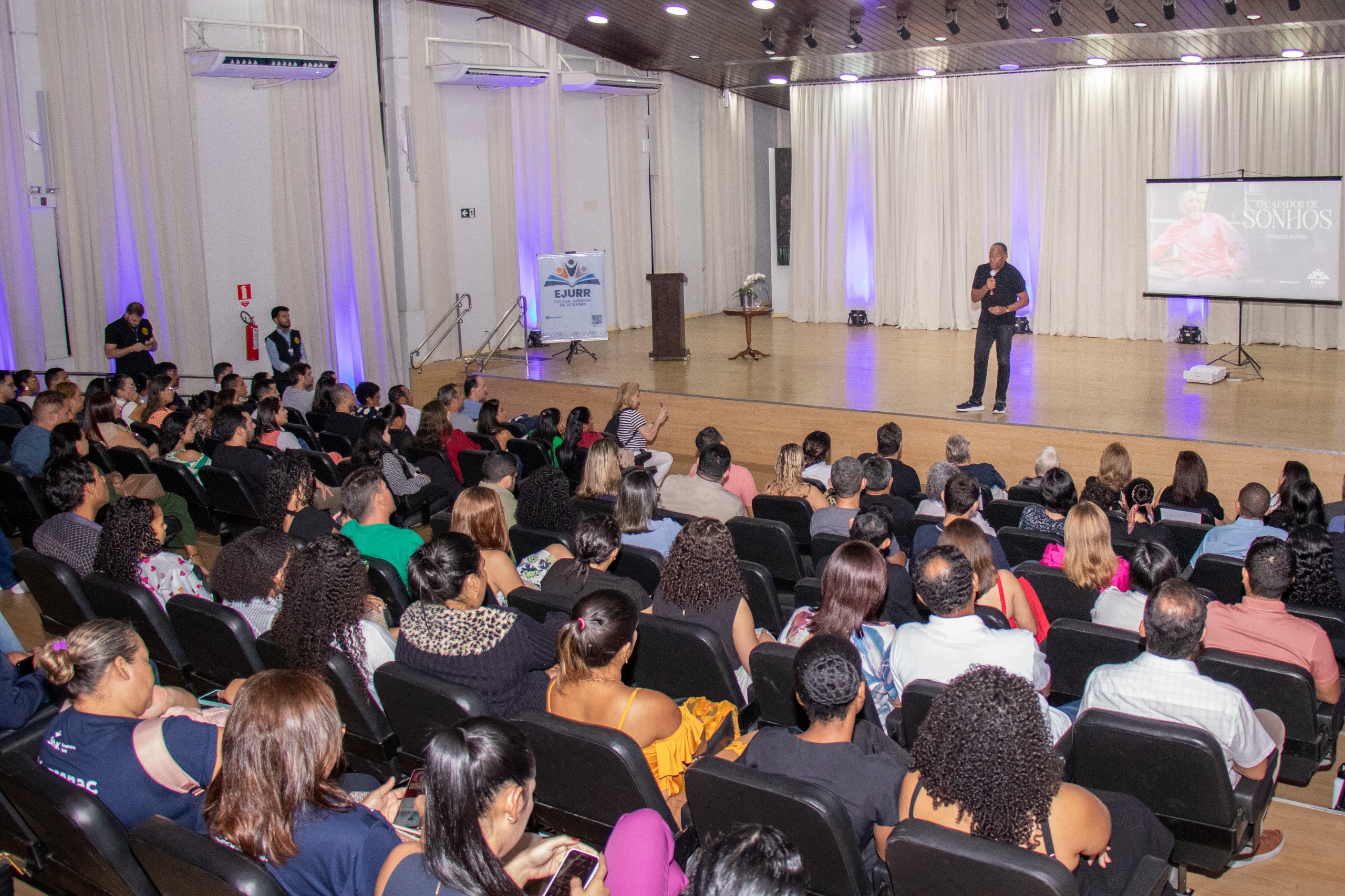 Imagem colorida mostrando o auditório do Fórum Cível cheio de pessoas durante palestra