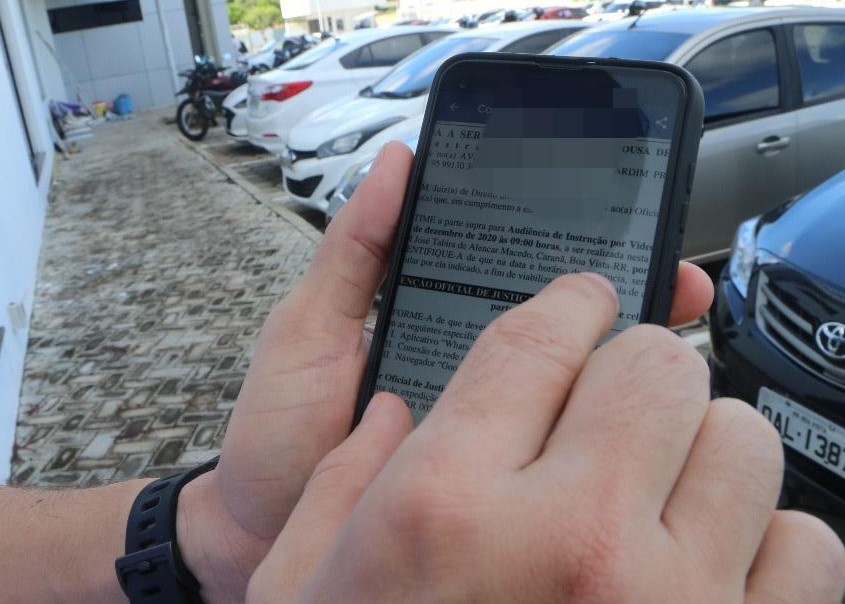 Foto de mãos de uma pessoa, segurando um telefone, em uma calçada com carros estacionados.