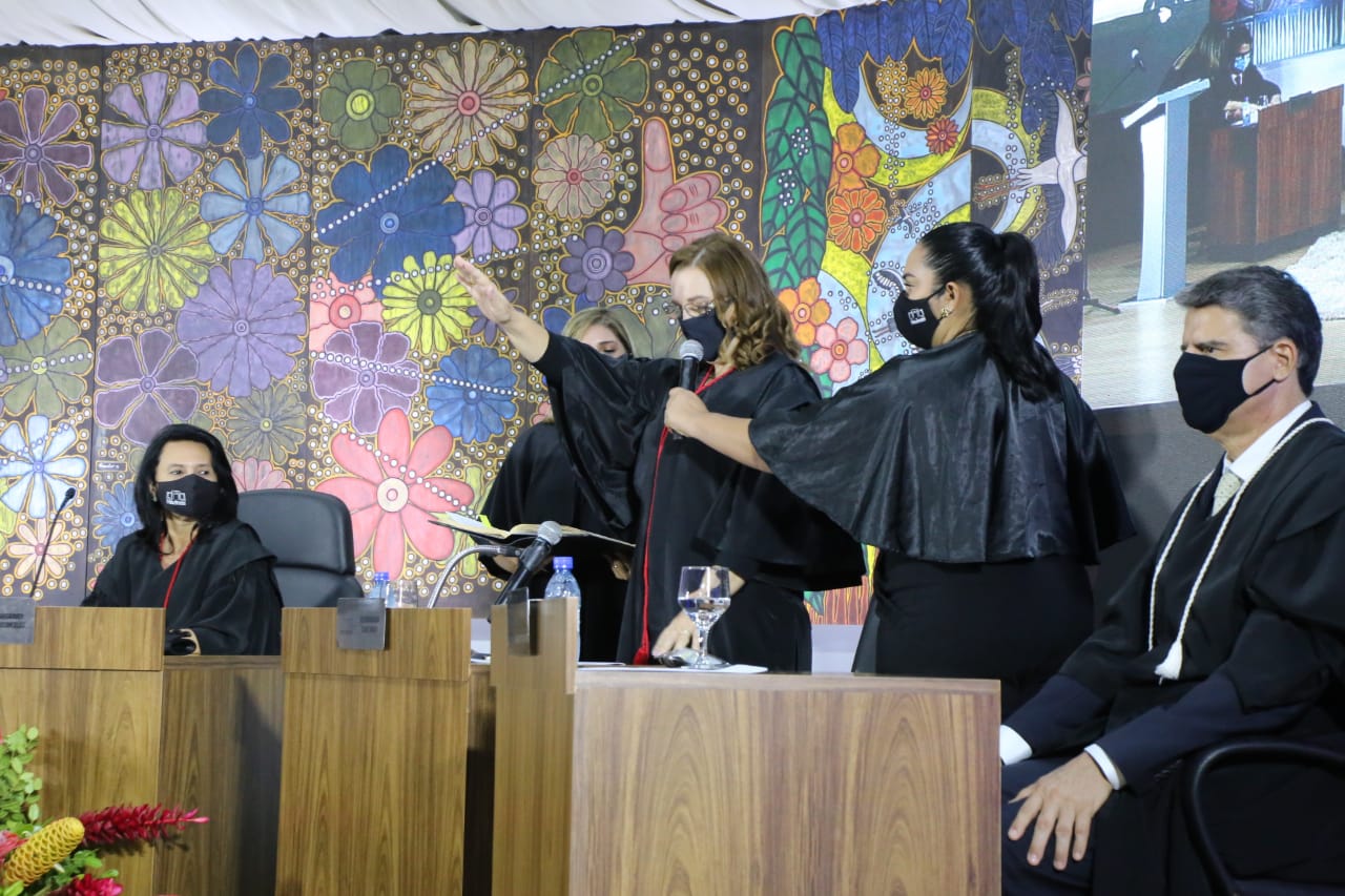 Foto de cerimônia de posse, ao centro a Desembargadora Elaine Bianchi, está de pé atrás da bancada, estendendo a mão e lendo um livro que está sendo segurado por uma mulher, que está de pé do lado esquerdo dela, ao lado direito uma outra mulher de pé segura o microfone que ela está falando. nas duas pontas da foto há dois magistrados sentados. O fundo é um painel colorido com imagens de flores.