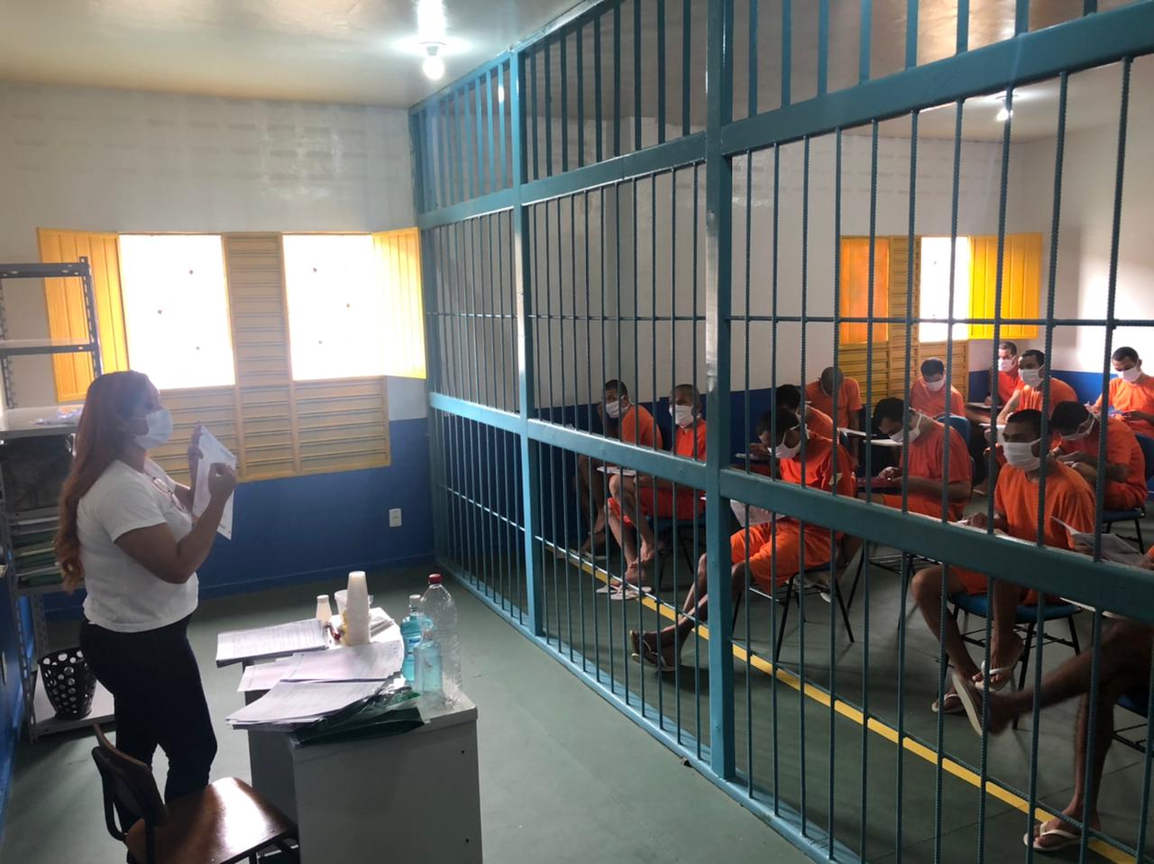 Foto de sala de aula dentro da penitenciária, ela é dividida por uma grade, de um lado a professora está de pé atrás de uma mesa com papéis, copos e garrafas, ao lado de uma cadeira e de um armário; e do outro lado da grade os detentos estão sentados em cadeiras, com papéis na mão e usando uniforme.