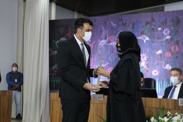 Magistrada de pé recebendo certificado de homem, ao fundo mesa com autoridades sentadas, em frente a um painel colorido, no canto esquerdo uma cortina e um servidor de pé.