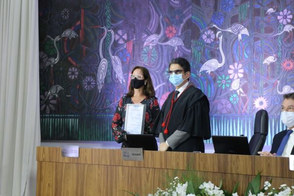 Foto do Desembargador Mozarildo Cavalcanti de pé, atrás da mesa do evento, ao lado de servidora que segura um certificado, ao lado deles, sentado está o Governador Antônio Denarium.