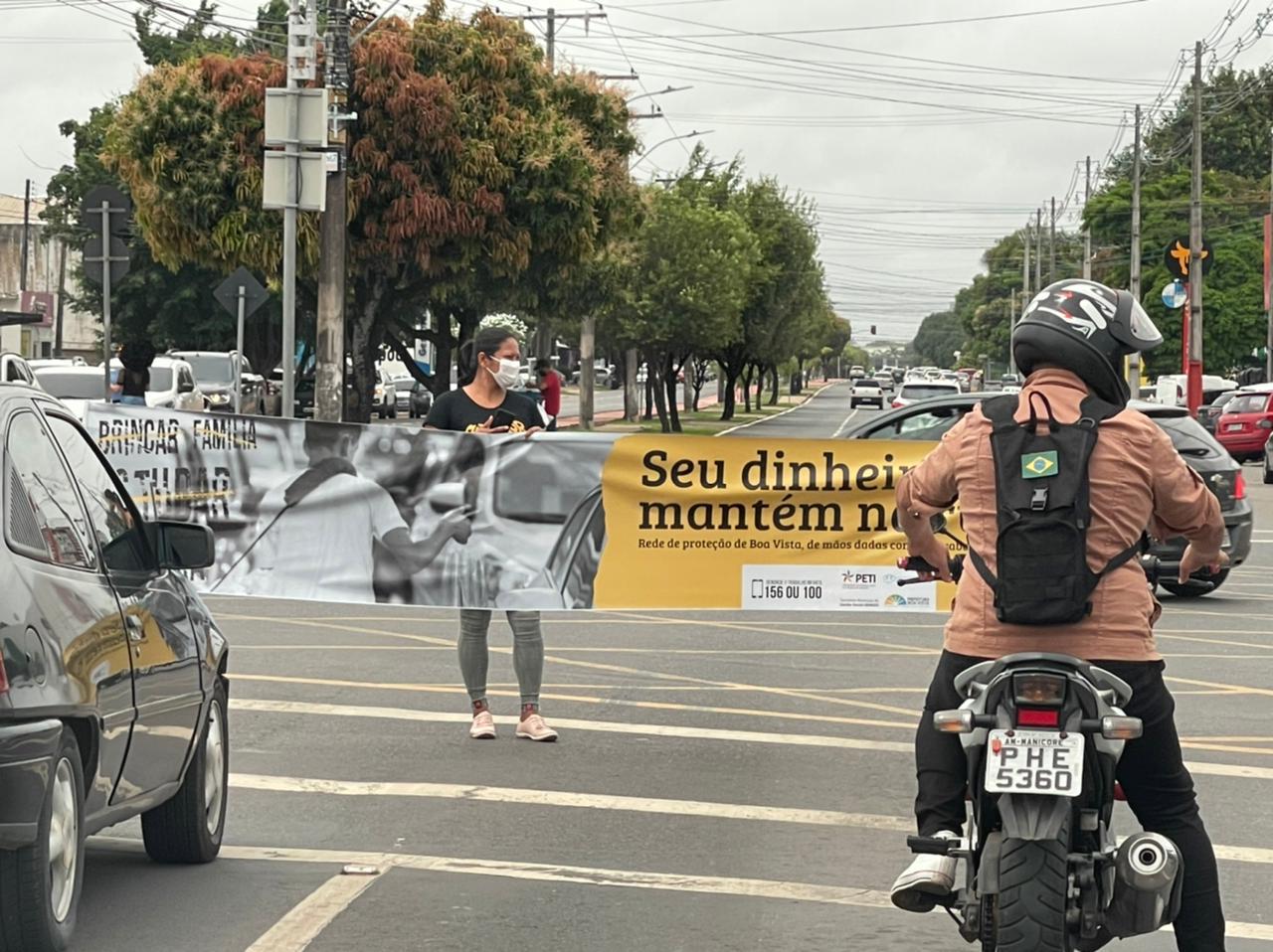 Foto de um carro e uma moto parados em um semáforo em uma avenida, na frente deles uma mulher segura uma faixa com a imagem de uma criança de costas, vendendo algo no sinal com a seguinte escrita: “Seu Dinheiro nos Mantém na Rua".