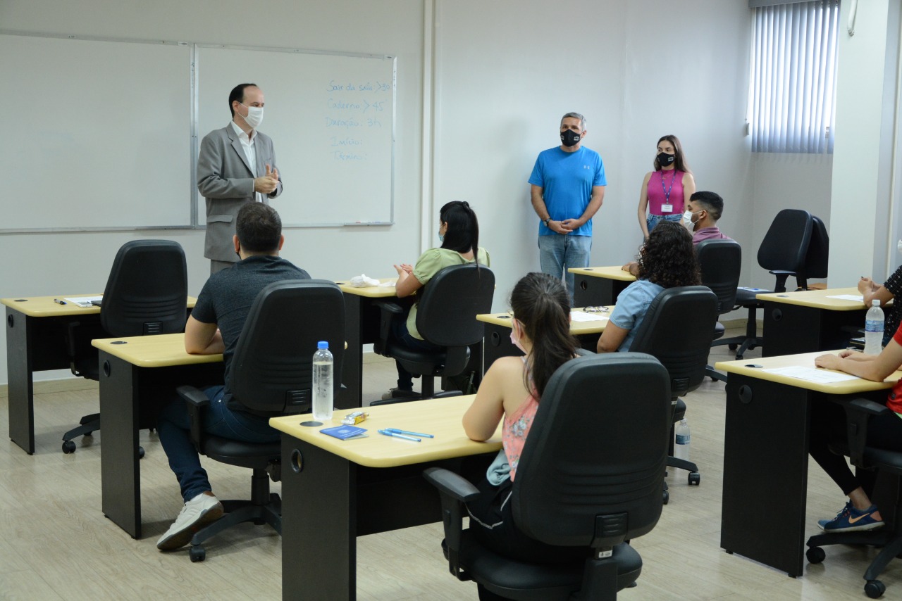 Em uma sala de aula, o presidente do TJRR, desembargador Cristóvão Suter, fala diante de candidatos. Ele é um homem de meia idade, alto, cabelos pretos, e está de terno bege, blusa e máscara brancas. Ele está acompanhado de um servidor e uma servidora do TJRR. 