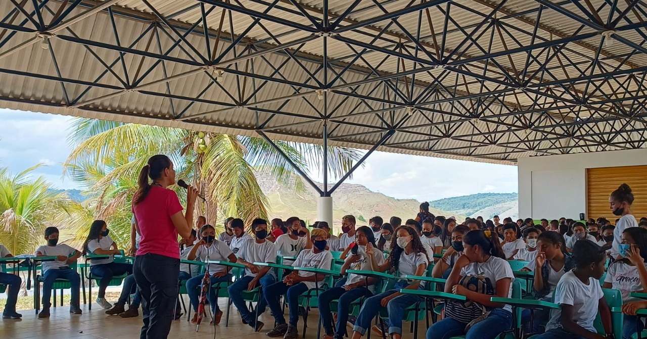 imagem colorida mostra ao lado direito, a palestrante Janaíne Voltolini de pé com um microfone na mão. À esquerda da foto estão alunos da Escola Estadual Joaquim Nabuco sentados.