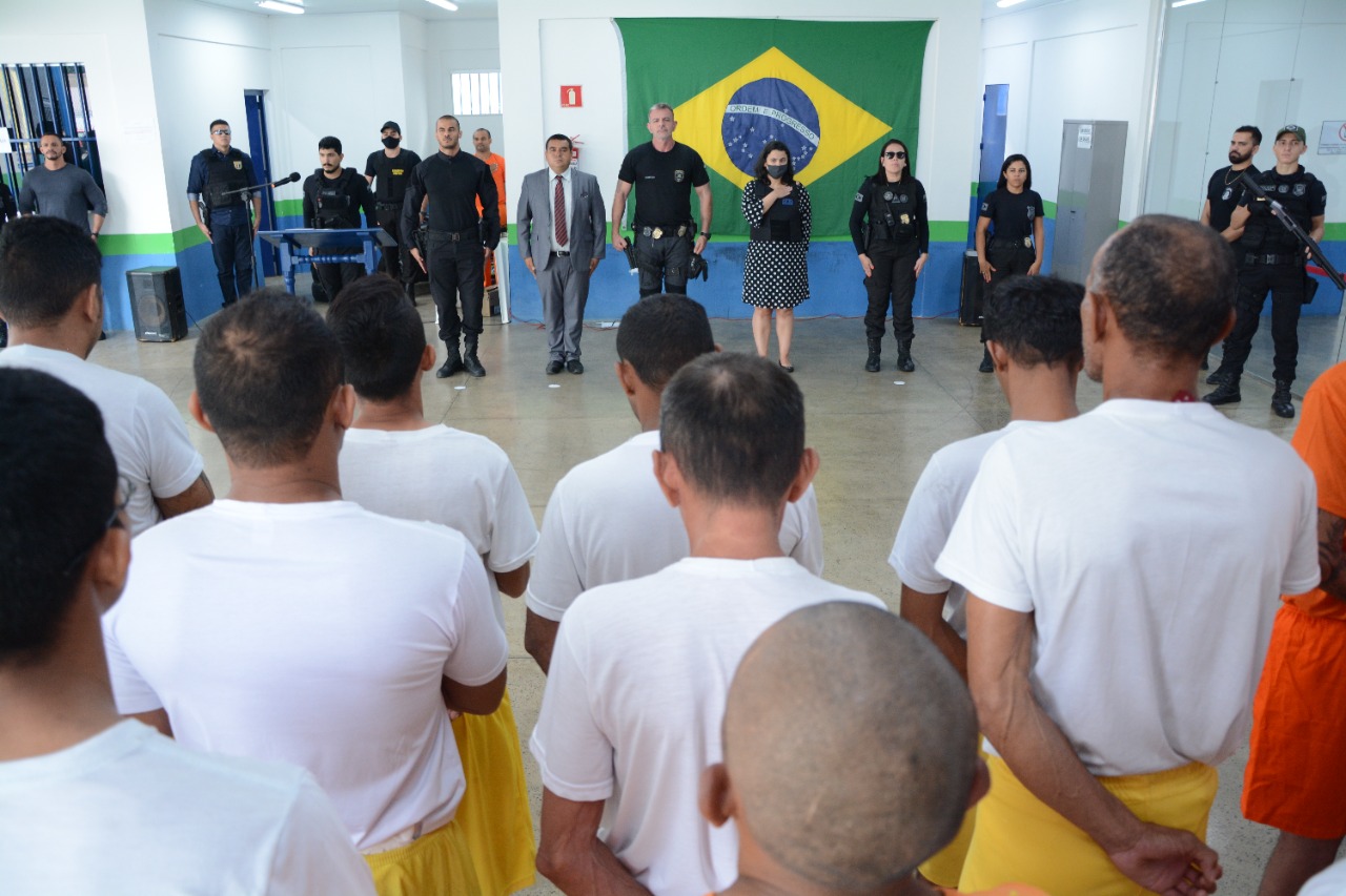 Imagem colorida durante solenidade na cadeia pública masculina, com os detentos de costas pra câmera e logo a frente deles, a juíza titular da Vara de Execução Penal do Tribunal de Justiça de Roraima (TJRR), Joana Sarmento, com a mão direita no coração, durante hino nacional brasileiro, juntamente com a equipe da unidade prisional.