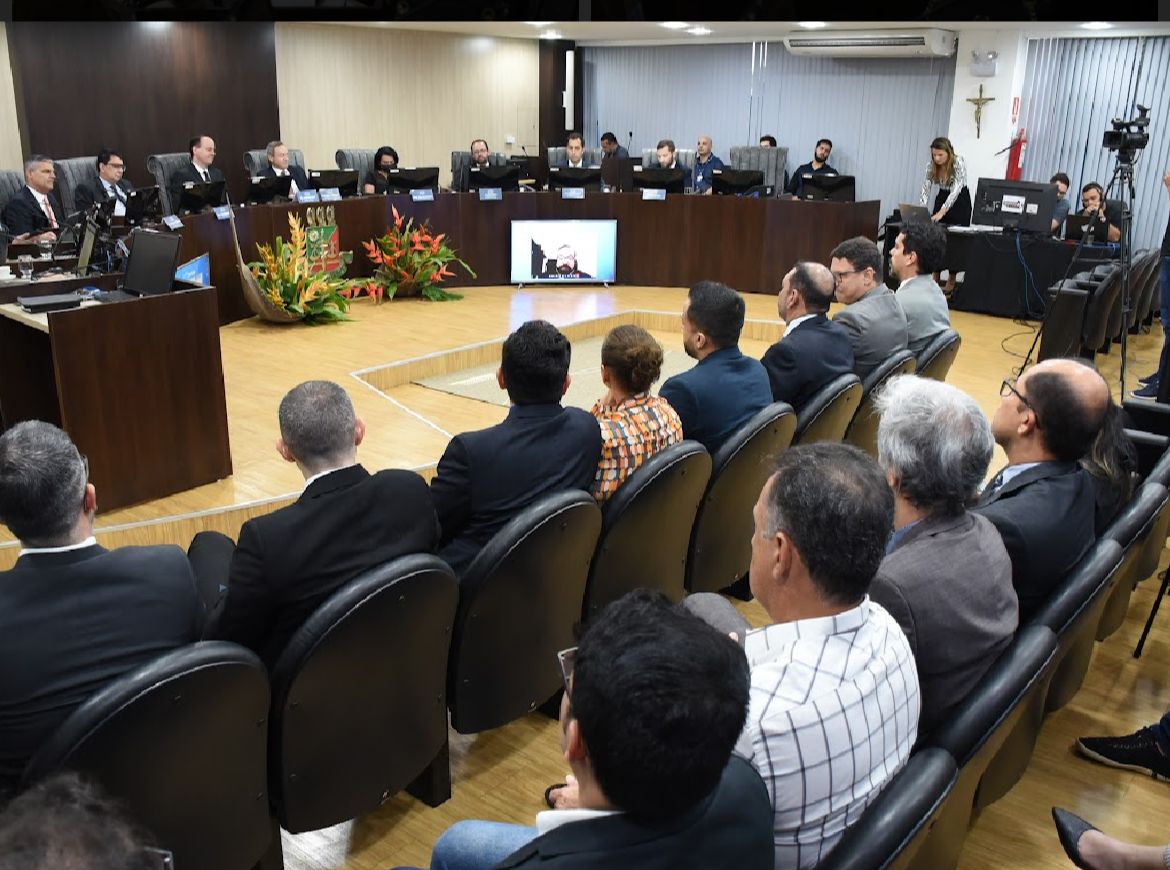 foto colorida mostra auditório do Tribunal Pleno com doze pessoas sentadas em poltronas pretas de frente para a tribuna. À frente estão oito magistrados sentados, em uma tribuna de cor marrom em formato de meia lua. Objetos como câmera, vasos de flores, computador e televisão aparecem na imagem.