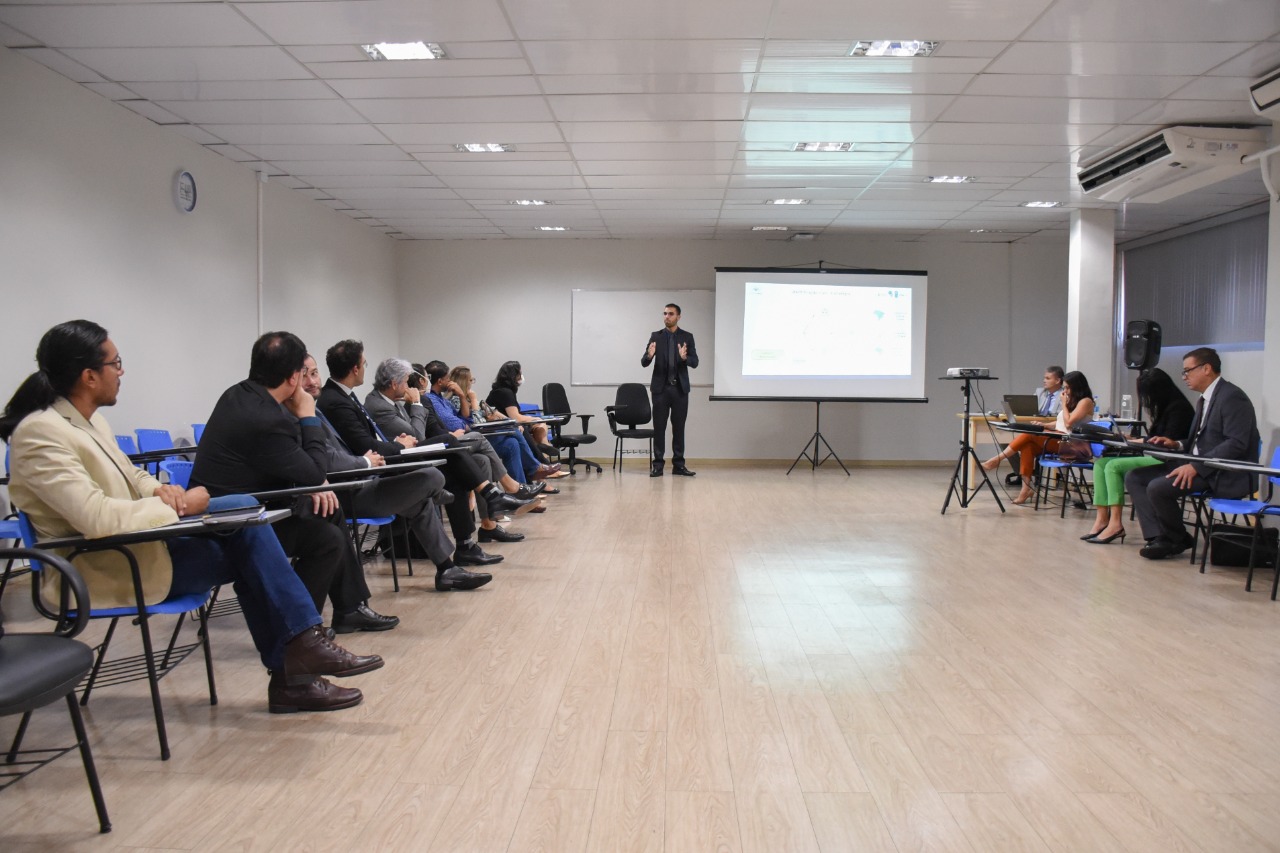 Foto quadrada colorida, com texto em destaque “Fazendo Justiça”. Imagem com nove pessoas sentadas em cadeiras azuis ao lado esquerdo. Ao centro está o coordenador do Programa das Nações Unidas para o Desenvolvimento do CNJ, em pé, gesticulando ao lado de um telão em que está sendo reproduzida uma imagem. Do lado direito da foto estão quatro pessoas sentadas em cadeiras azuis. Todas estão observando a fala do coordenador.