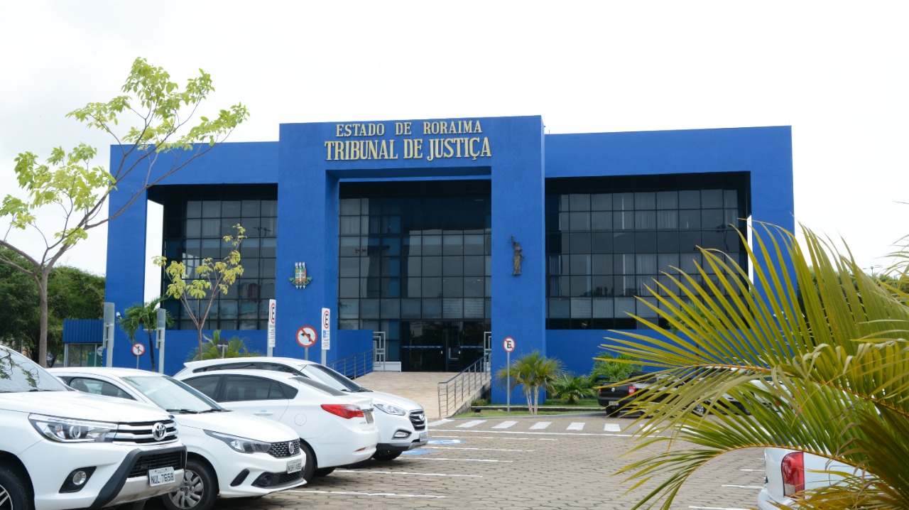 A imagem colorida contém fachada do Palácio da Justiça vista pelo ângulo do estacionamento que fica em frente a entrada do palácio. Na imagem é possível também ver carros estacionados enfileirados e uma parte do jardim que fica em frente a entrada. 