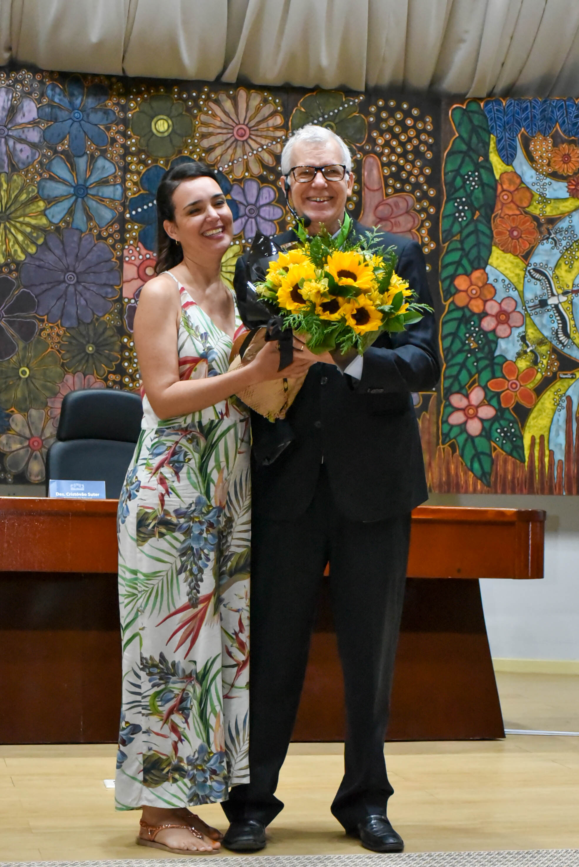 Imagem colorida contém o ministro do STJ, recebendo das mãos da servidora do TJRR, Rudianna Zeidler, um buquê de flores.