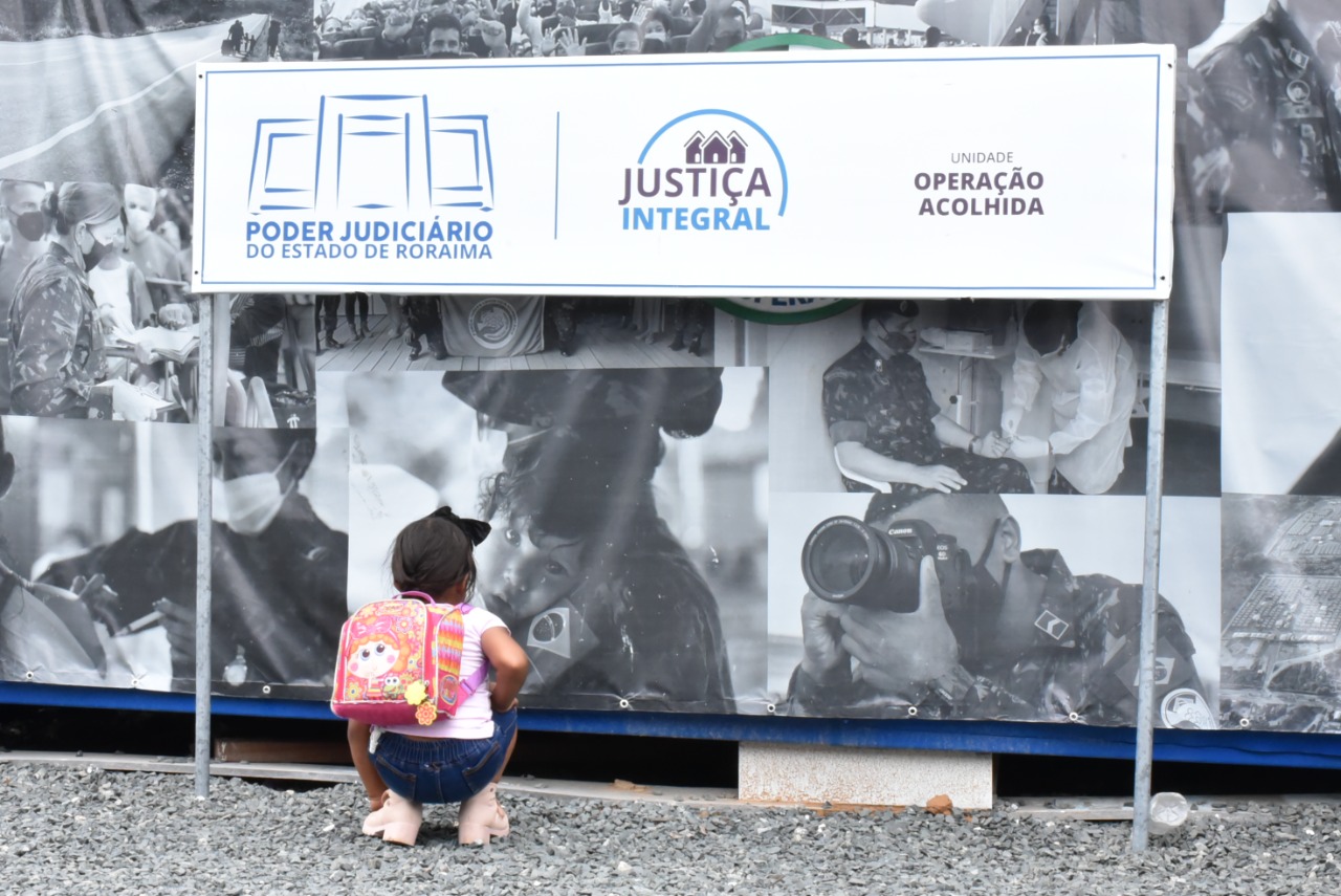  Imagem colorida contém criança olhando para placa do polo do Justiça Integral. localizado no  localizado no Posto de Triagem e Interiorização da operação Acolhida. 