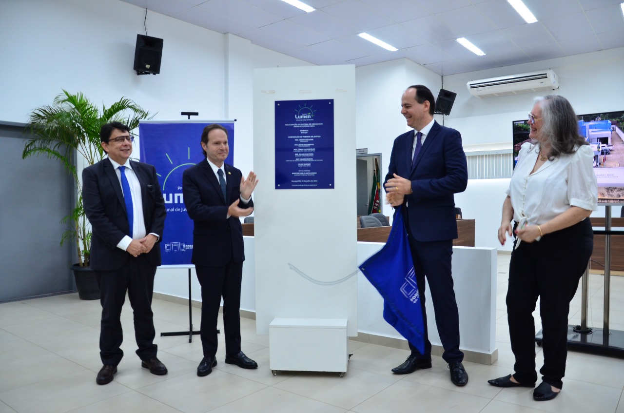  Imagem colorida contém uma placa do projeto lumen à esquerda da placa está dois homens um está rindo e o outro aplaudindo, do lado direito da placa está um homem segurando uma bandeira azul e ao seu lado está uma mulher sorridente.