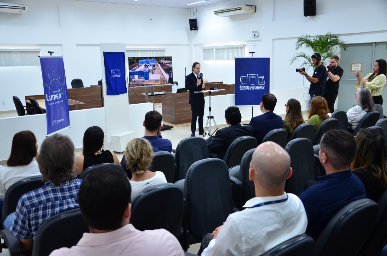 Imagem colorida de um auditório com plateia e um facilitador discursando para  público presente.