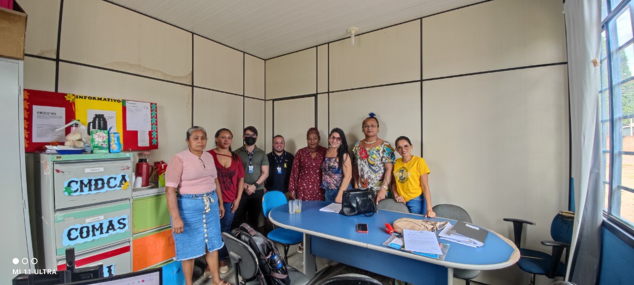 Imagem colorida em formato retangular mostra uma sala com 7 pessoas em pé, uma mesa azul no centro e sobre ela há papéis e bolsas, na lateral esquerda da imagem armários coloridos e um quadro de mensagens