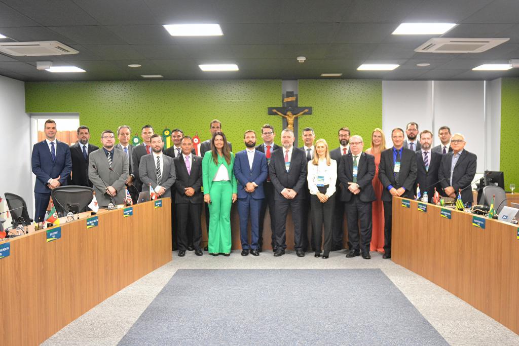 Imagem colorida mostra o juiz auxiliar da Presidência e a coordenadora do Núcleo de Precatórios do Tribunal de Justiça de Roraima, Valdira Silva, com os  desembargadores, juízes e assessores, de todos os estados brasileiros em pé próximos a uma  bancada com bandeiras de cada estado.