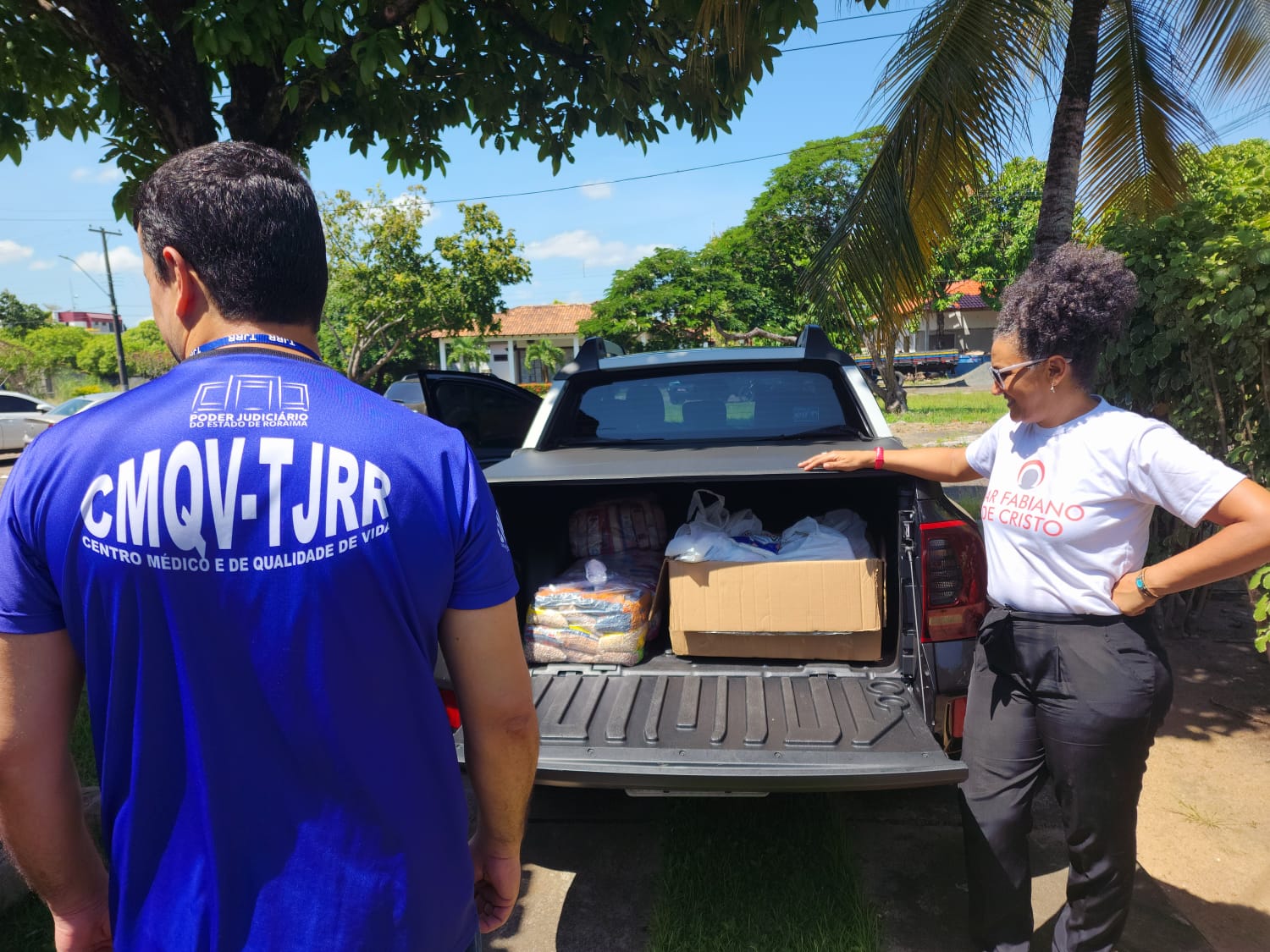 Imagem colorida mostra uma Picape Strada com caixas e sacolas de alimentos, uma mulher encostando no carro e um funcionário do TJRR com a camisa do Centro Médico e de Qualidade de Vida.