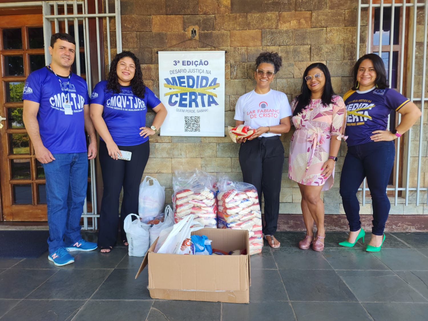 Imagem colorida mostra caixas, sacos e sacolas com alimentos, à esquerda da foto estão dois funcionários do TJRR, e à direita estão três funcionárias do Lar Fabiano de Cristo.