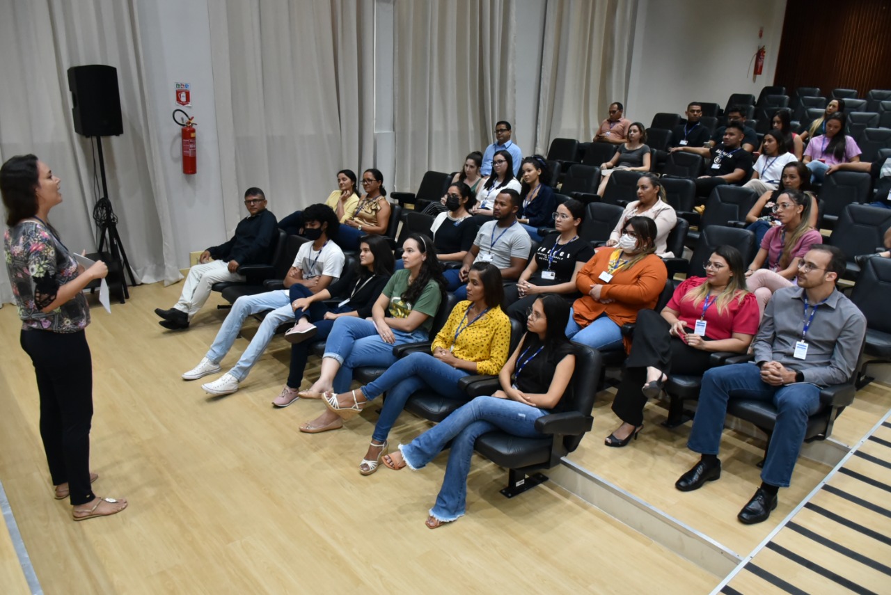 Imagem colorida mostra a servidora Rudianna Zeigler palestrando em um auditório para um pequeno público.