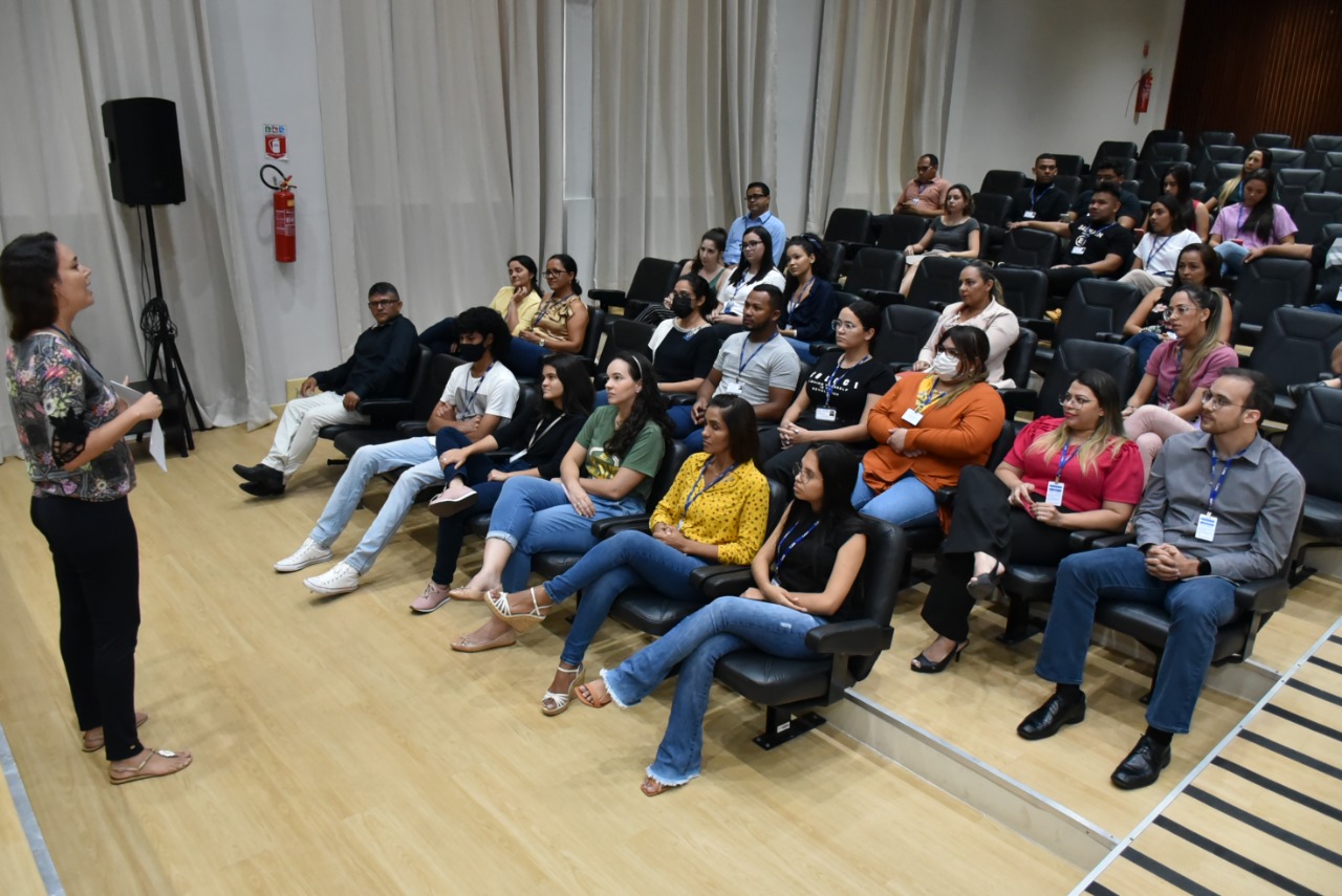 Imagem colorida mostra a servidora Rudianna Zeigler palestrando em um auditório para um pequeno público. 