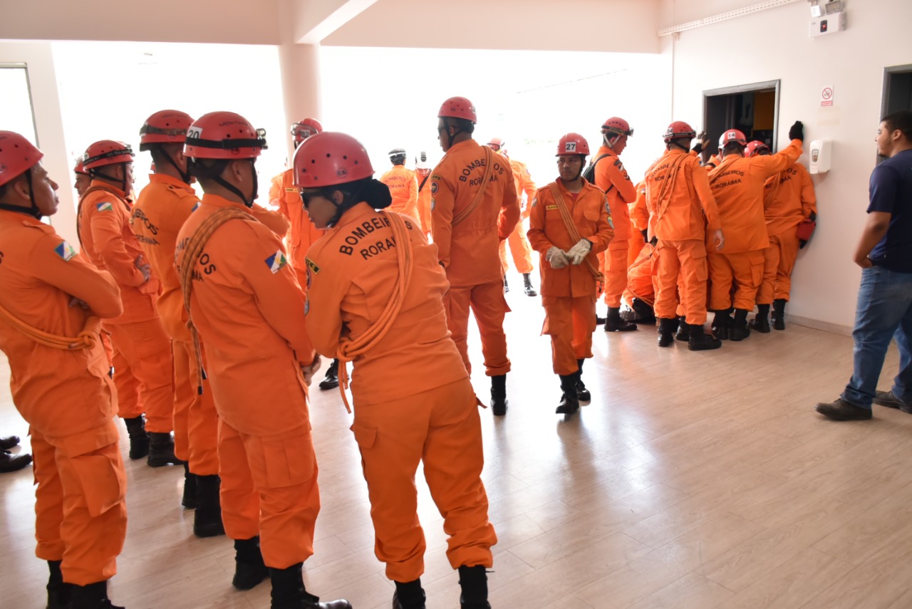 Imagem colorida mostra 26 alunos Cabos com capacetes e roupas no tom laranja em pé em uma sala.