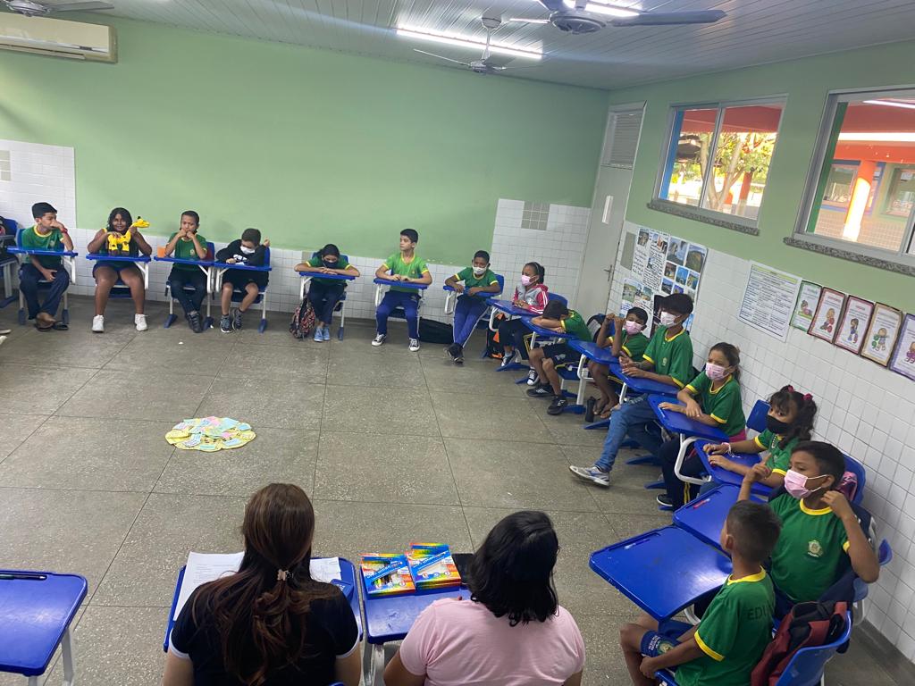 Imagem colorida contém crianças em sala de aula, sentadas durante roda de conversa com duas pedagogas do Programa Maria Vai à Escola do TJRR.