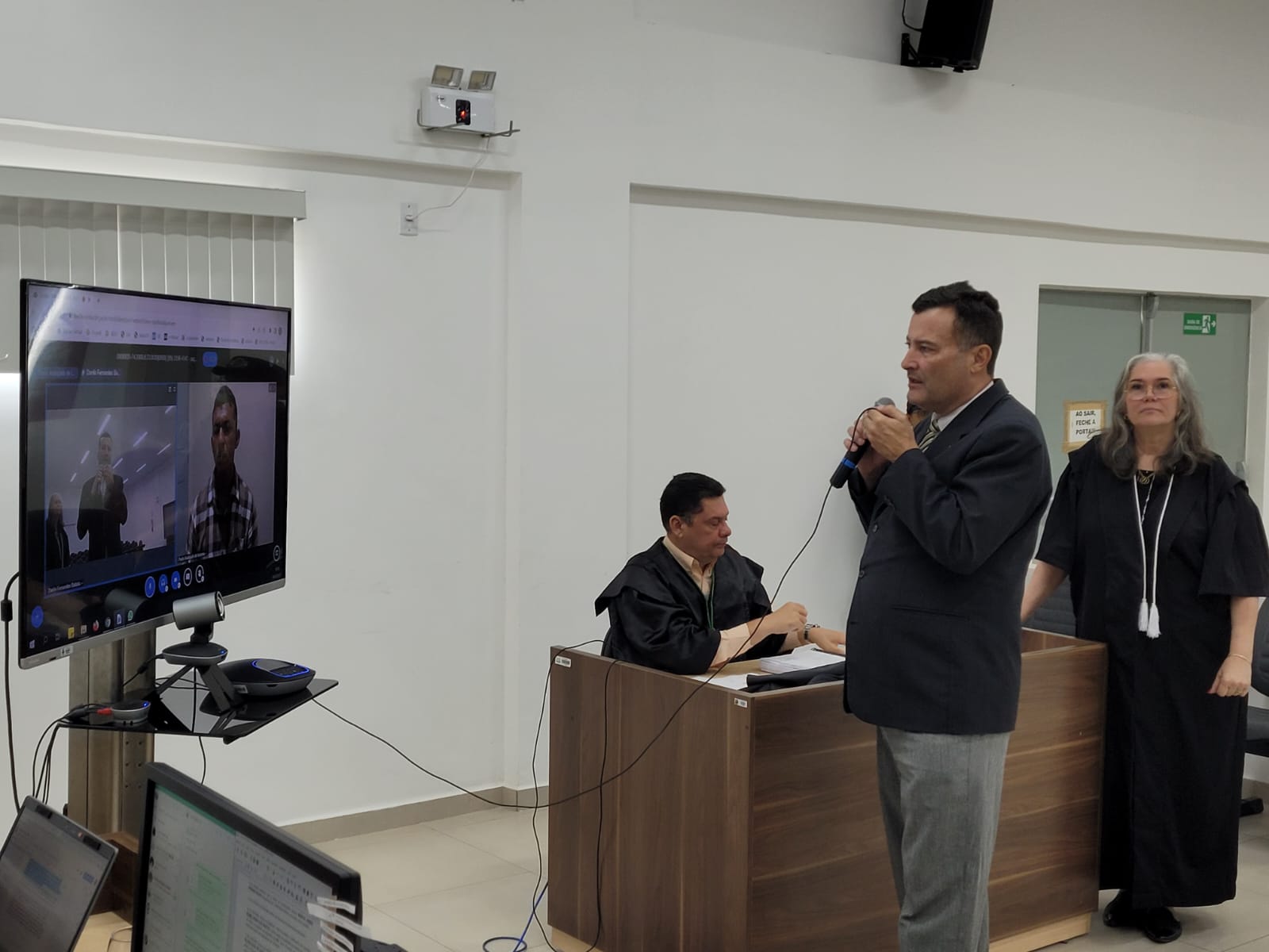 Imagem colorida contém três pessoas no mutirão de sessão do Tribunal do Júri, na comarca de Mucajaí. Um homem se encontra de frente para o televisor segurando o microfone, tendo atrás dele uma juíza em pé e o outro juiz sentado. 