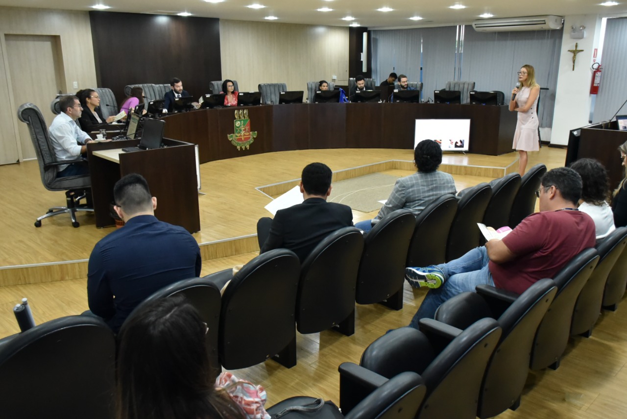 Imagem colorida contém servidores do TJRR no Tribunal do Pleno durante concurso do Prêmio de Inovação do Poder Judiciário. Ao fundo da imagem encontram-se a banca de qualificação do concurso e a chefe do Setor de Enfrentamento à Violência Doméstica do TJRR apresentando. 