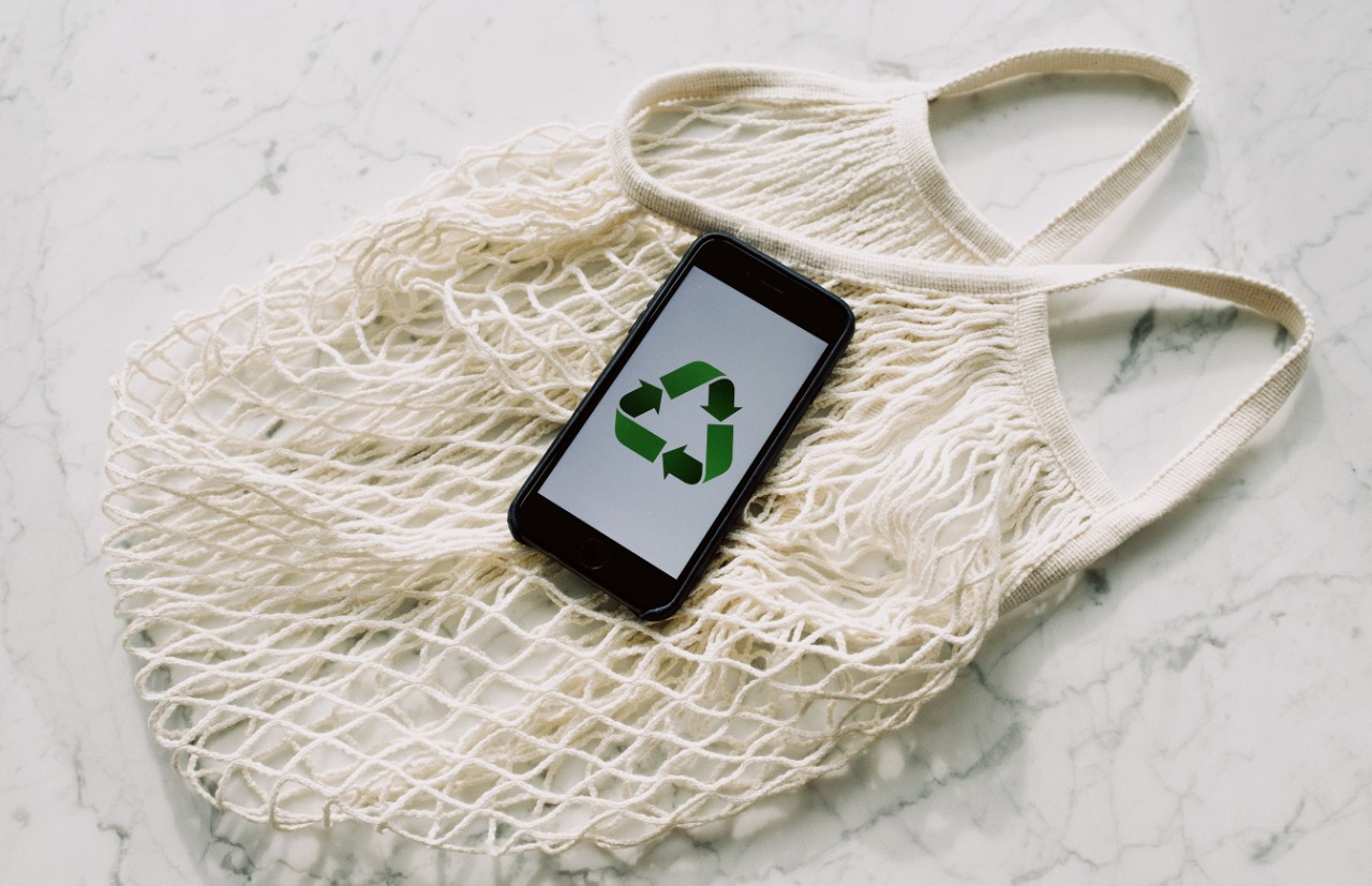 Imagem colorida ilustrativa mostra um celular com o símbolo de reciclagem na tela, o mesmo encontra-se em cima de uma bolsa de saco branca que está sobre de uma bancada de mármore. 