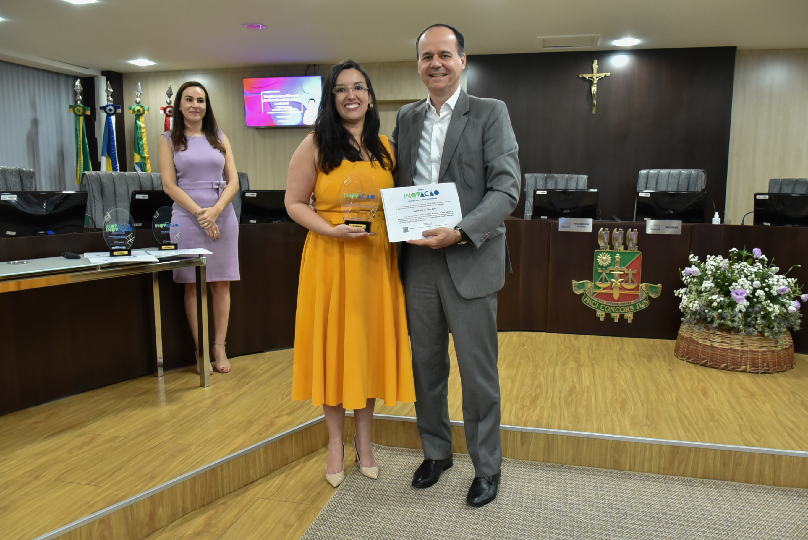 Imagem colorida em formato retangular mostra o presidente Cristóvão Suter segurando o certificado do prêmio inovação ao seu lado está com  a juíza Suelen Alves, a mesma  segura o troféu da premiação. 