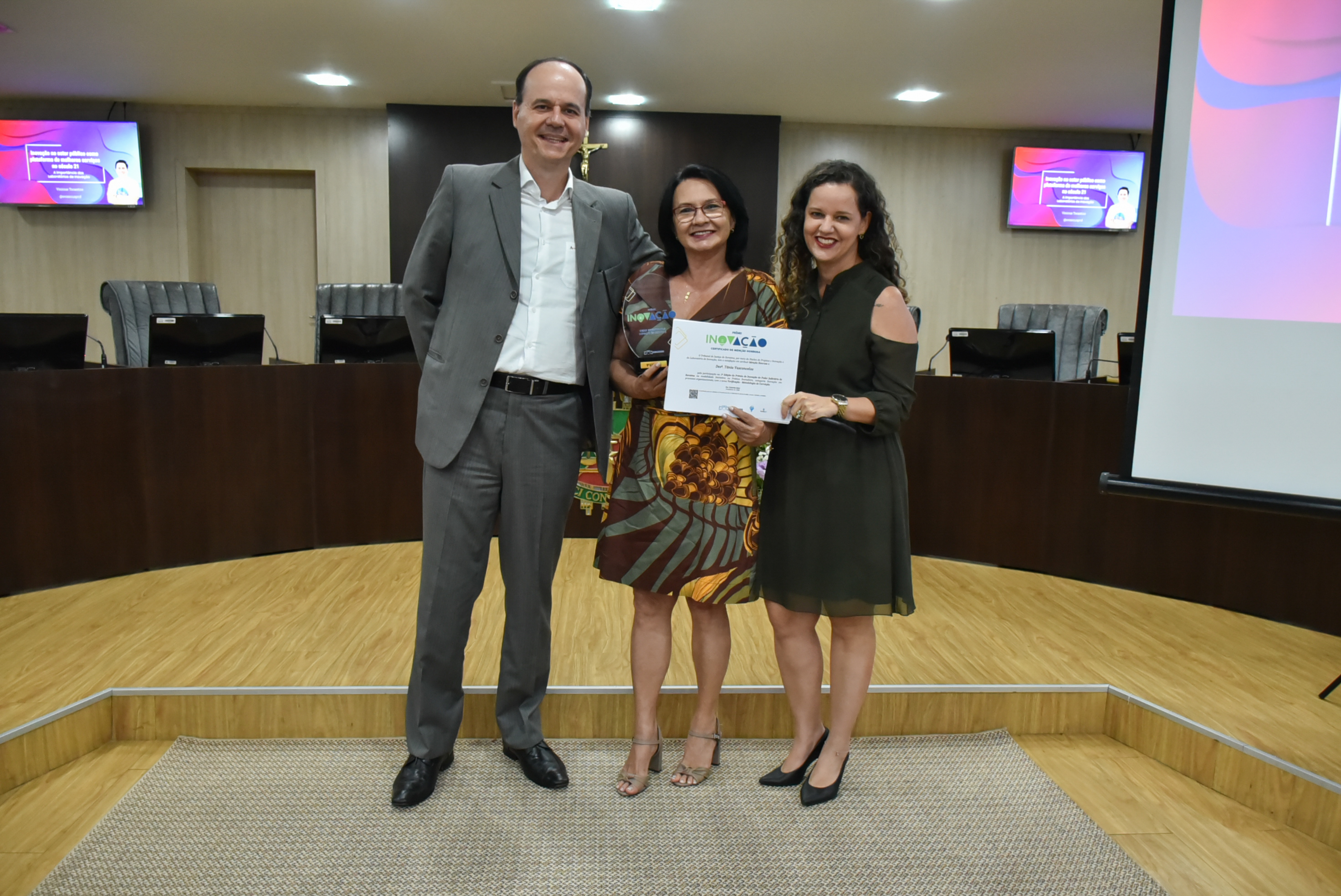  Imagem colorida em formato retangular mostra o presidente Cristóvão Suter ao lado da   desembargadora Dra. Tânia Vasconcelos que segura em suas mãos o troféu e o certificado junto com a diretora de Gestão do 1º Grau, Inaiara Sá.