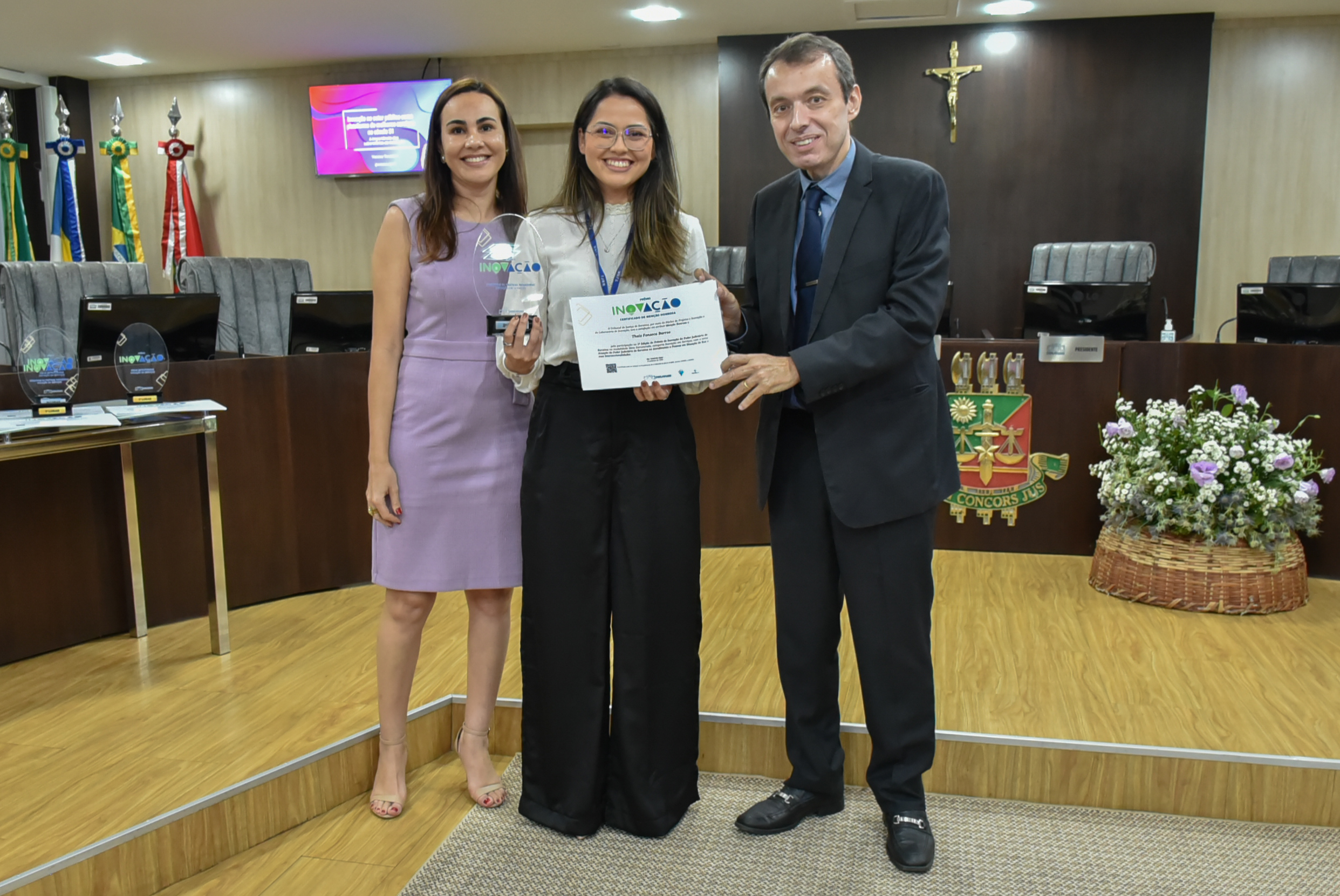  Imagem colorida em formato retangular mostra servidora no centro da foto segurando o troféu e o certificado do prêmio inovação,  acompanhada do desembargador Erick Linhares e da coordenadora do Núcleo de Projetos e Inovação, Janaine Voltolin.