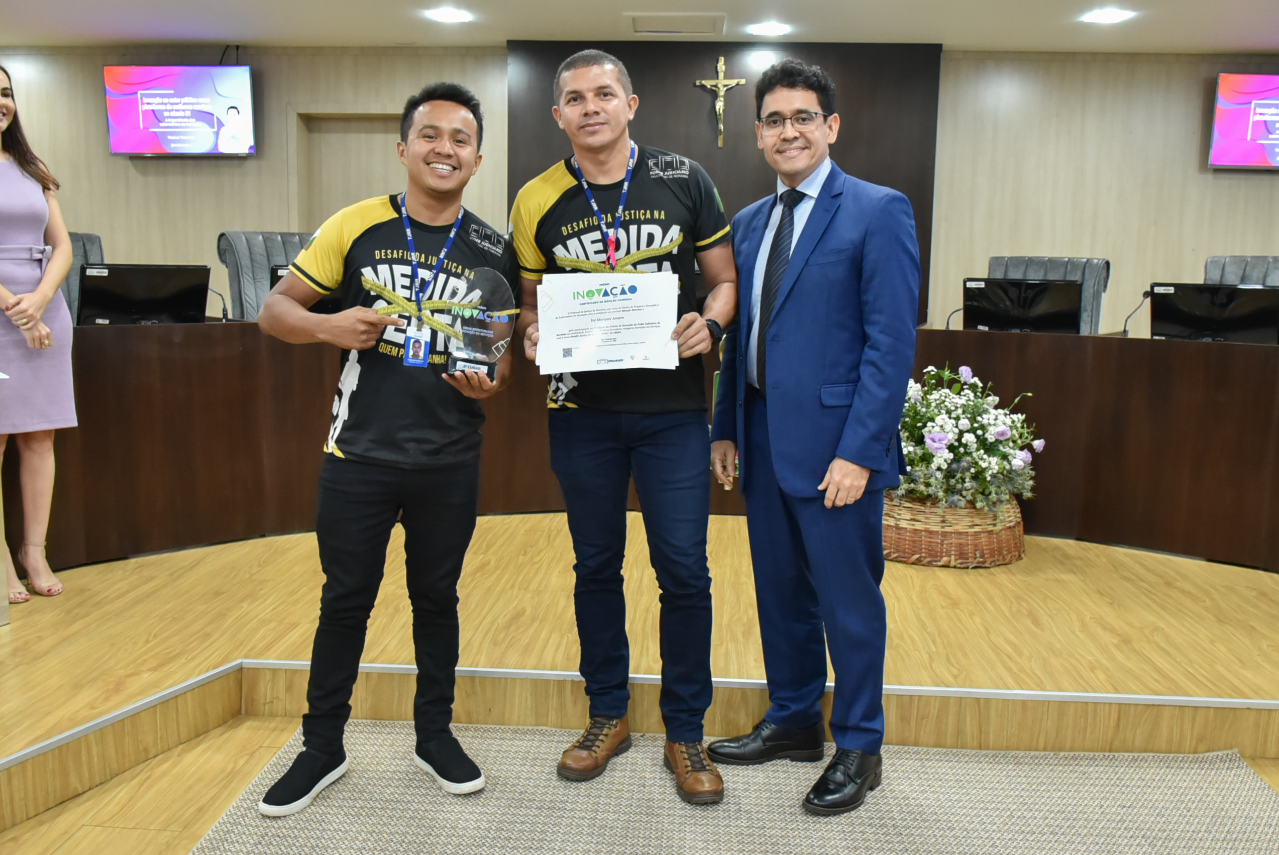 Imagem colorida em formato retangular mostra os educadores físicos do TJRR,  Ronaldo e Marcio Cavalcante, segurando o troféu e o certificado do prêmio inovação,  acompanhados do juiz  auxiliar da Corregedoria de Justiça  do Poder Judiciário, Breno Coutinho.