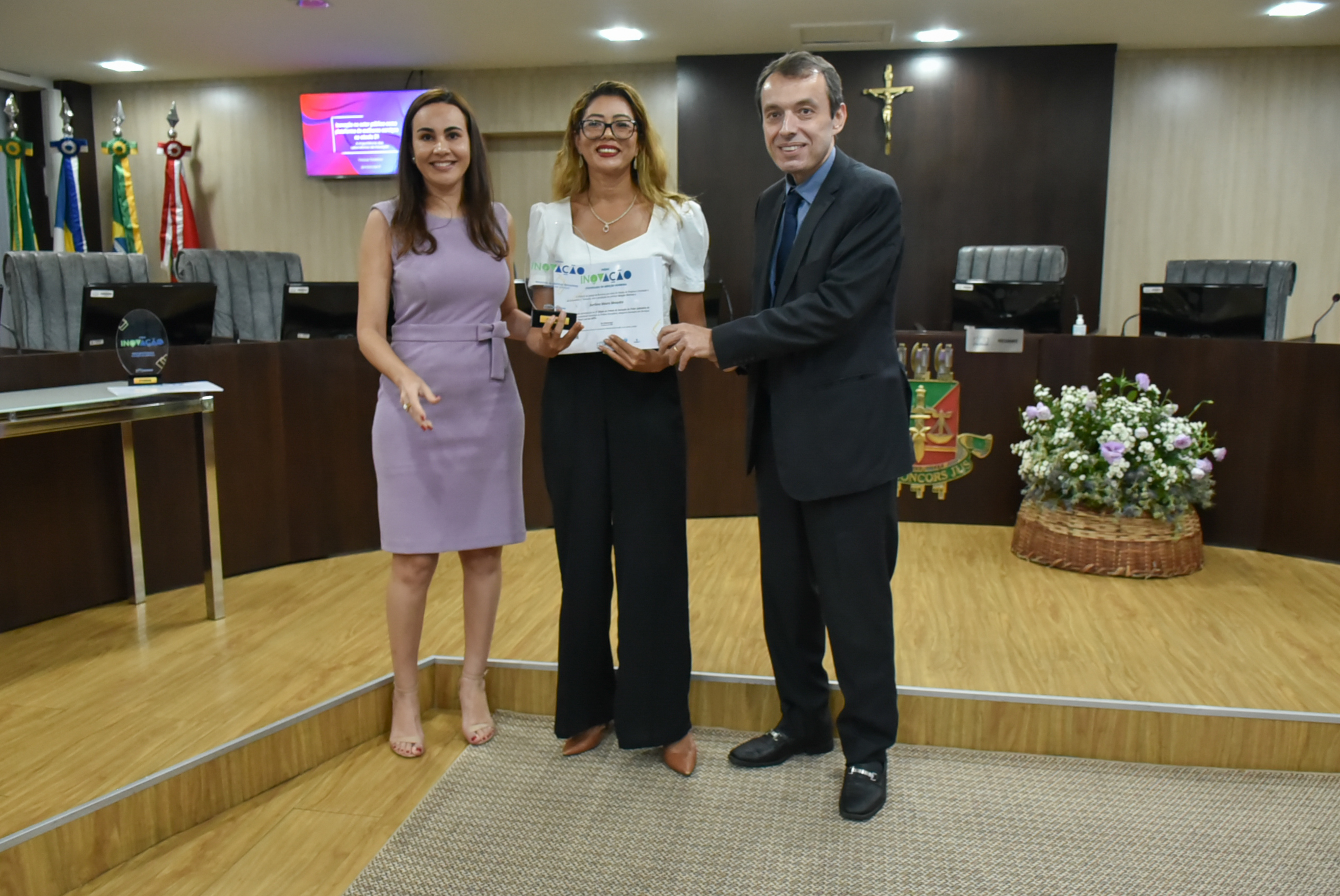 Imagem colorida em formato retangular mostra a chefe do Setor de Enfrentamento à Violência Doméstica, Aurilene Mesquita no centro da foto segurando o troféu e o certificado do prêmio inovação,  acompanhada do desembargador Erick Linhares. e da coordenadora do Núcleo de Projetos e Inovação, Janaine Voltolin.