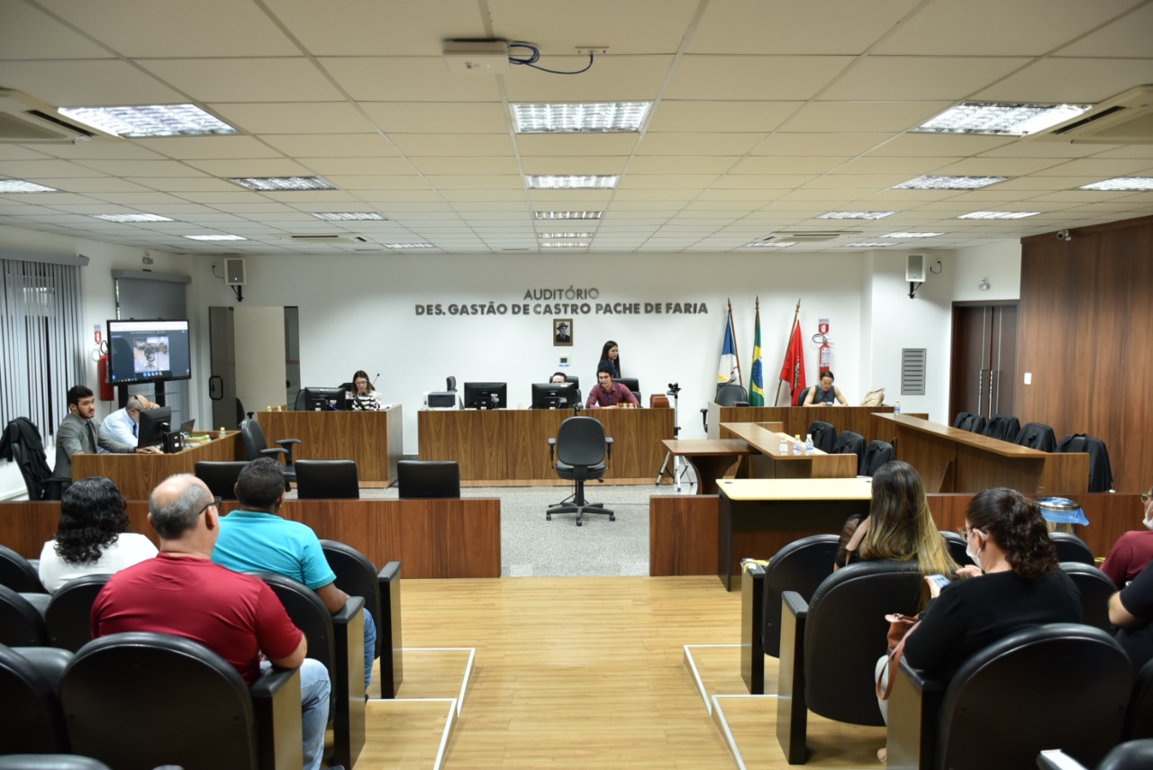 Imagem colorida em formato retangular mostra uma audiência no Fórum Criminal Ministro Evandro Lins e Silva com algumas pessoas reunidas, entre elas há homens e mulheres. Na sala há assentos pretos e bancadas na cor marfim, em cima das bancadas há computadores. No fundo da imagem há 3 bandeiras hasteadas.