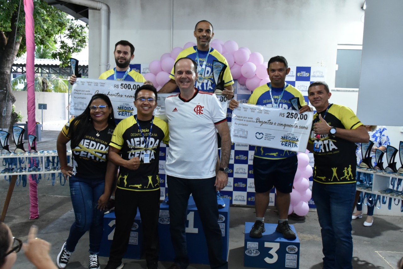 imagem colorida mostra o presidente do TJRR, desembargador Cristóvão Suter, acompanhado dos três vencedores da categoria masculina de perda de percentual de gordura corporal e dos educadores físicos do TJ. Os vencedores encontram-se em cima do podium com suas medalhas e com um cheque representando o valor de sua premiação.