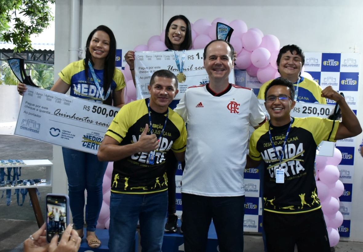 imagem colorida mostra o presidente do TJRR, desembargador Cristóvão Suter, acompanhado das três vencedoras da categoria feminina de perda de percentual de gordura corporal e dos educadores físicos do TJ. As vencedoras encontram-se em cima do podium com suas medalhas e com um cheque representando o valor de sua premiação.