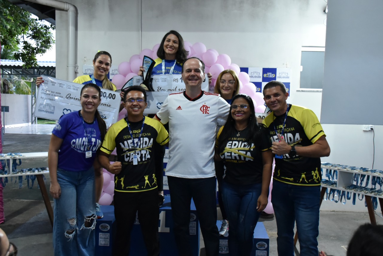 imagem colorida mostra o presidente do TJRR, desembargador Cristóvão Suter, acompanhado das três vencedoras da categoria feminina de ganho percentual de massa muscular esquelética e dos educadores físicos do TJ. As vencedoras encontram-se em cima do podium com suas medalhas e com um cheque representando o valor de sua premiação.