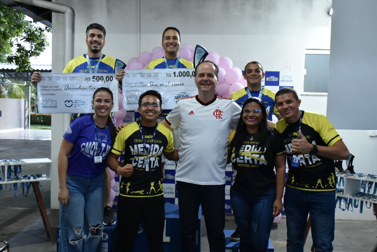imagem colorida mostra o presidente do TJRR, desembargador Cristóvão Suter, acompanhado dos três vencedores da categoria masculina de ganho percentual de massa muscular esquelética e dos educadores físicos do TJ. Os vencedores encontram-se em cima do podium com suas medalhas e com um cheque representando o valor de sua premiação.